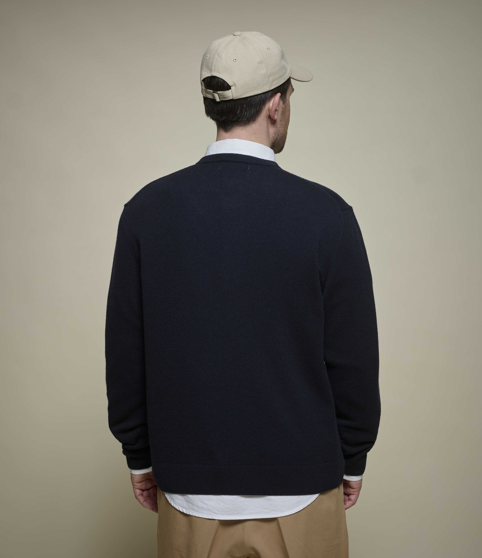 A man viewed from behind wearing a dark navy blue knit henley sweater over a white collared shirt, khaki pants, and a beige baseball cap, set against a plain beige background.