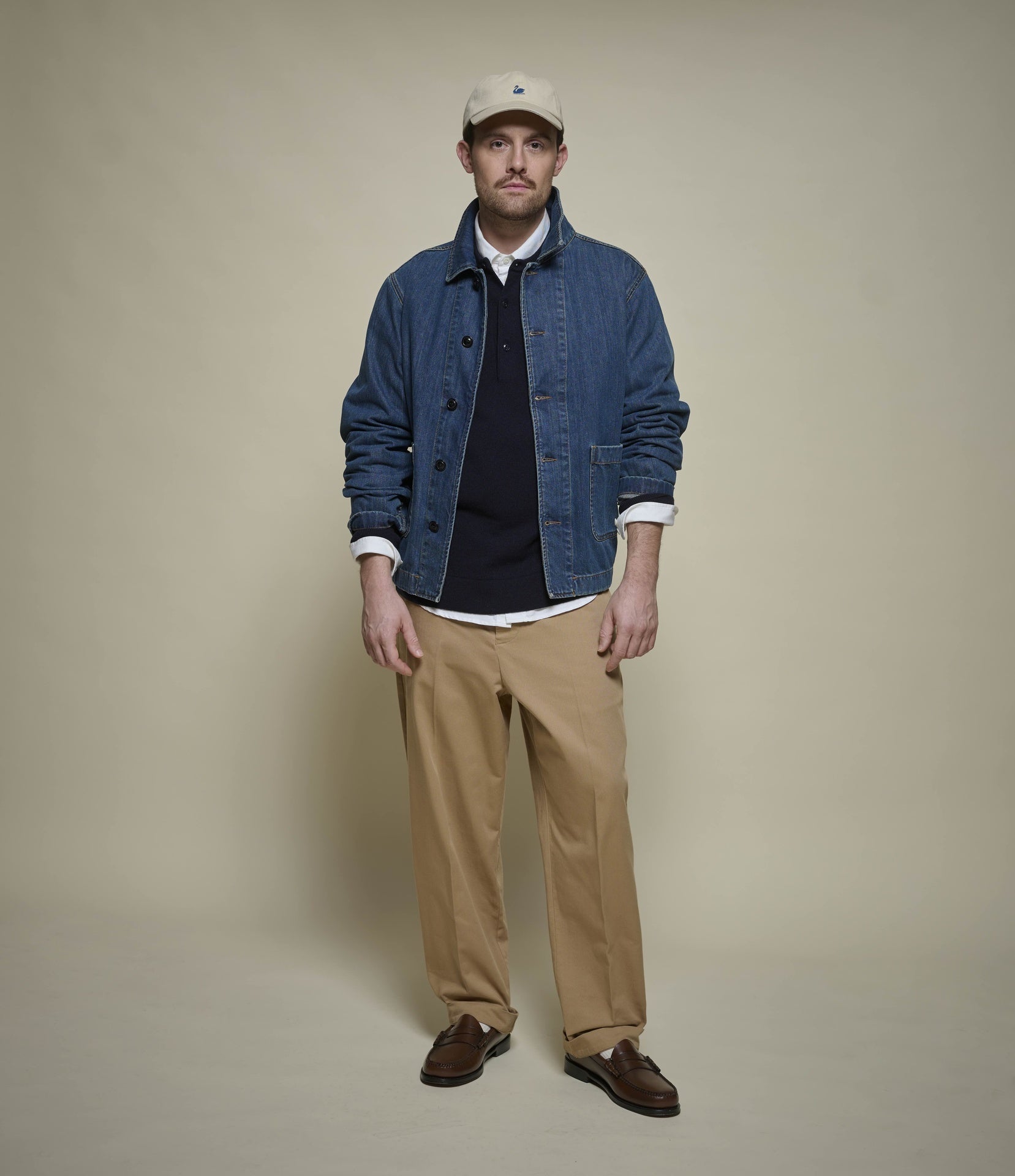 A man modeling a blue denim jacket over a blue henley shirt, white collared shirt, tan pants, and brown shoes, standing against a plain beige studio background.