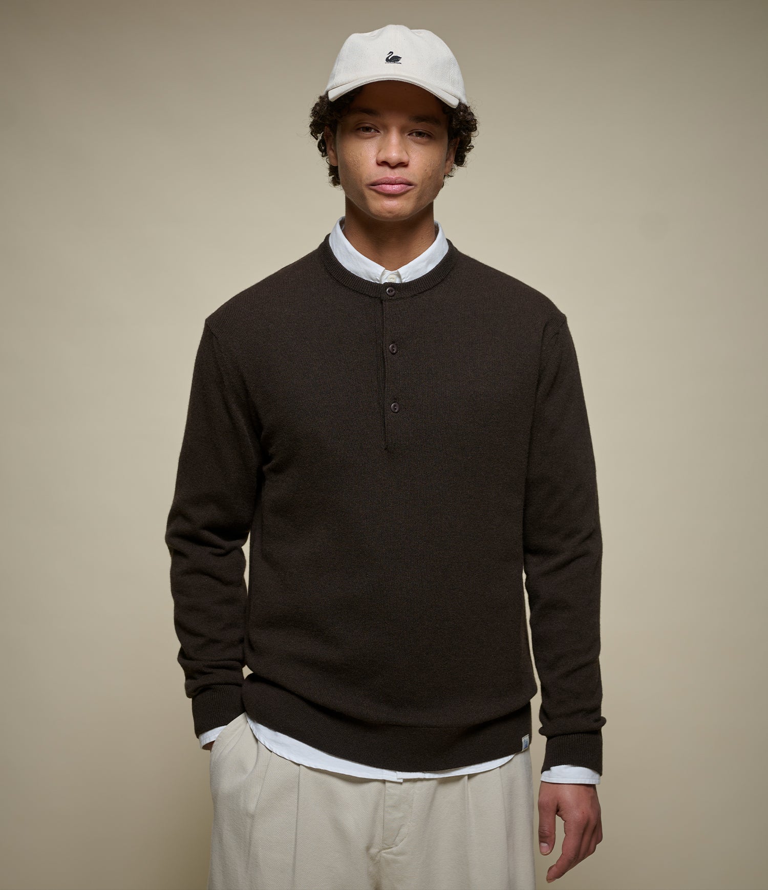 A man wearing a dark brown long-sleeve henley sweater over a white collared shirt, paired with light beige trousers and a white baseball cap, standing against a solid beige background.