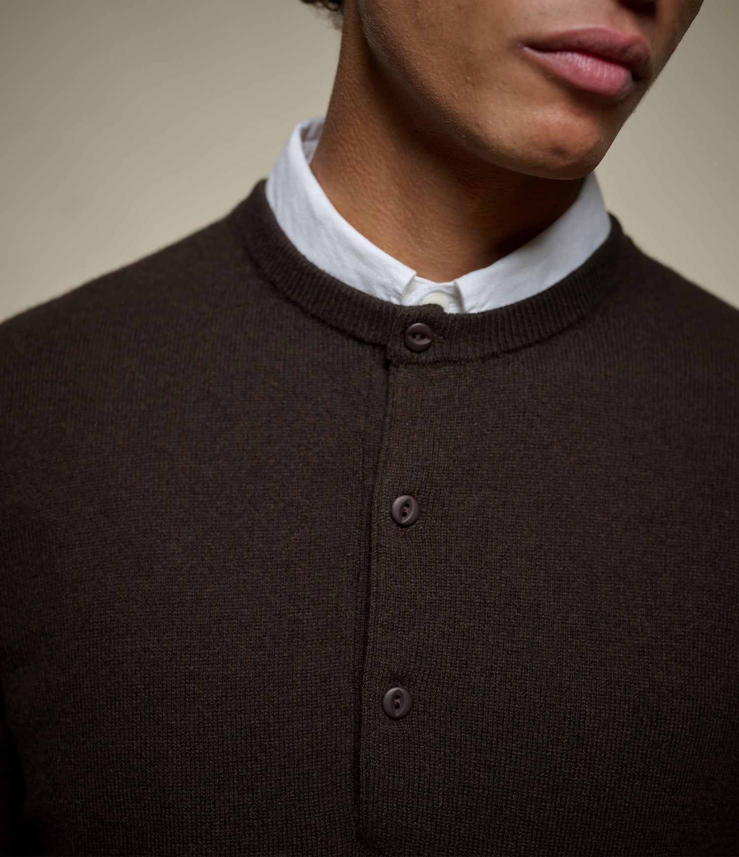 Close-up of a person wearing a dark brown ribbed knit henley sweater over a white collared shirt, against a plain beige background.