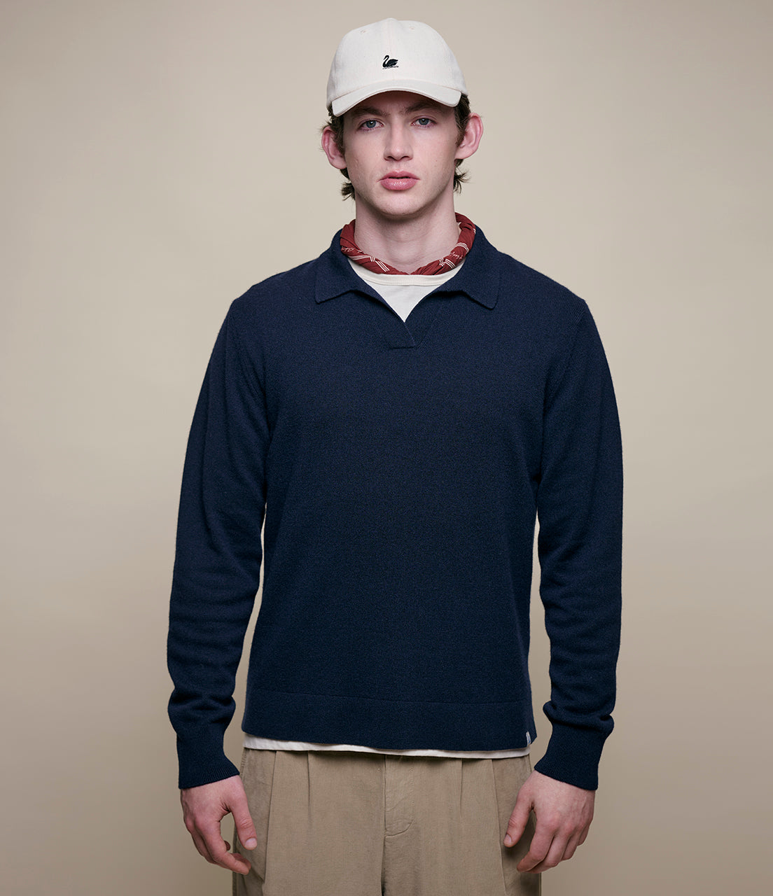 Man wearing a navy long-sleeve shirt, beige pants, and a white cap with a logo on a beige background