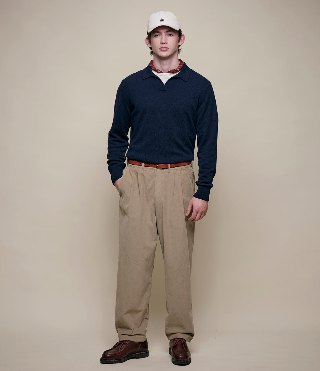Man wearing a navy sweater, beige pants, and a white cap against a beige background