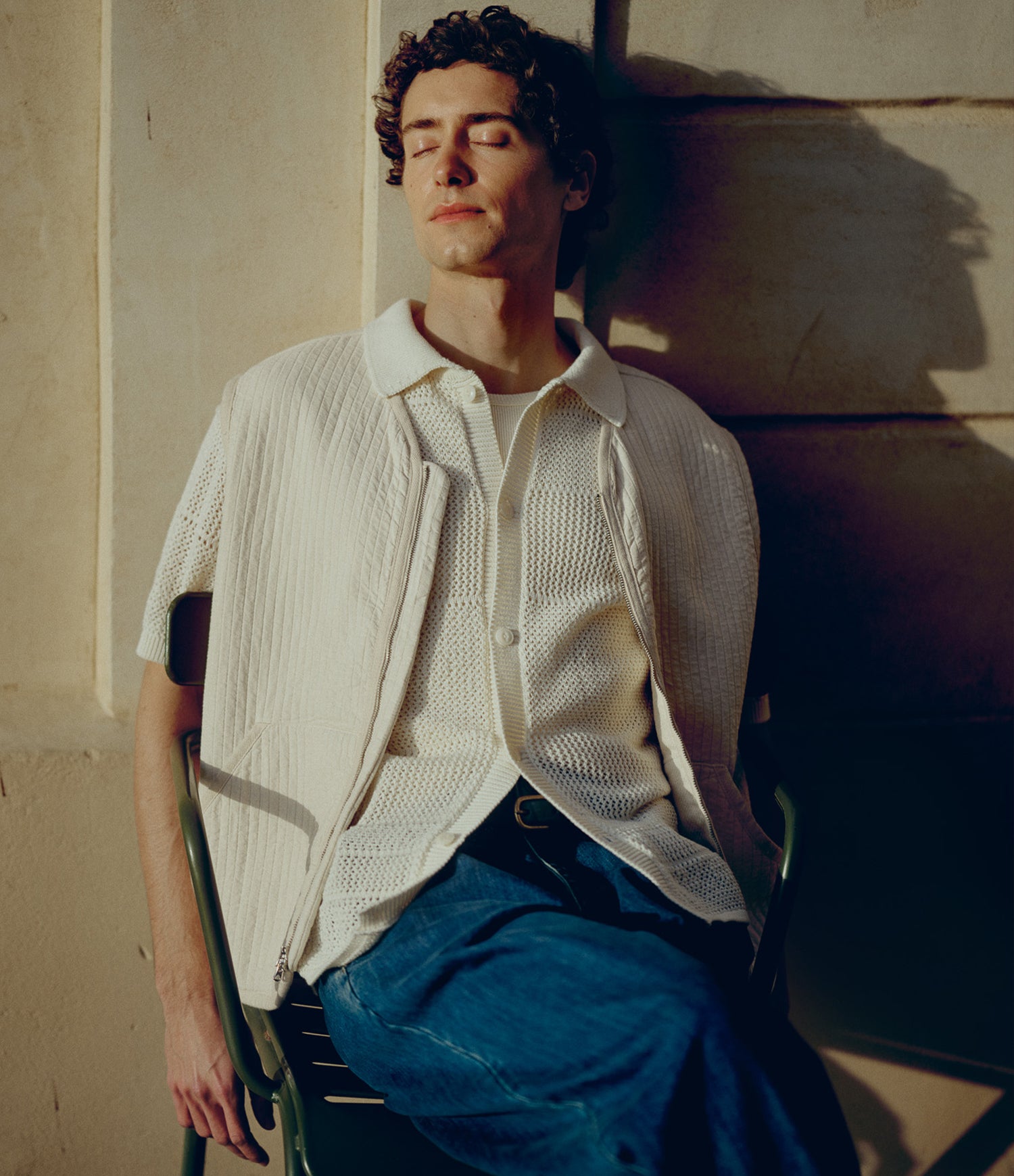 A man reclining on a chair against a sunlit stone wall, wearing a cream textured short-sleeve button-front overshirt over a matching cream knit top with blue jeans.
