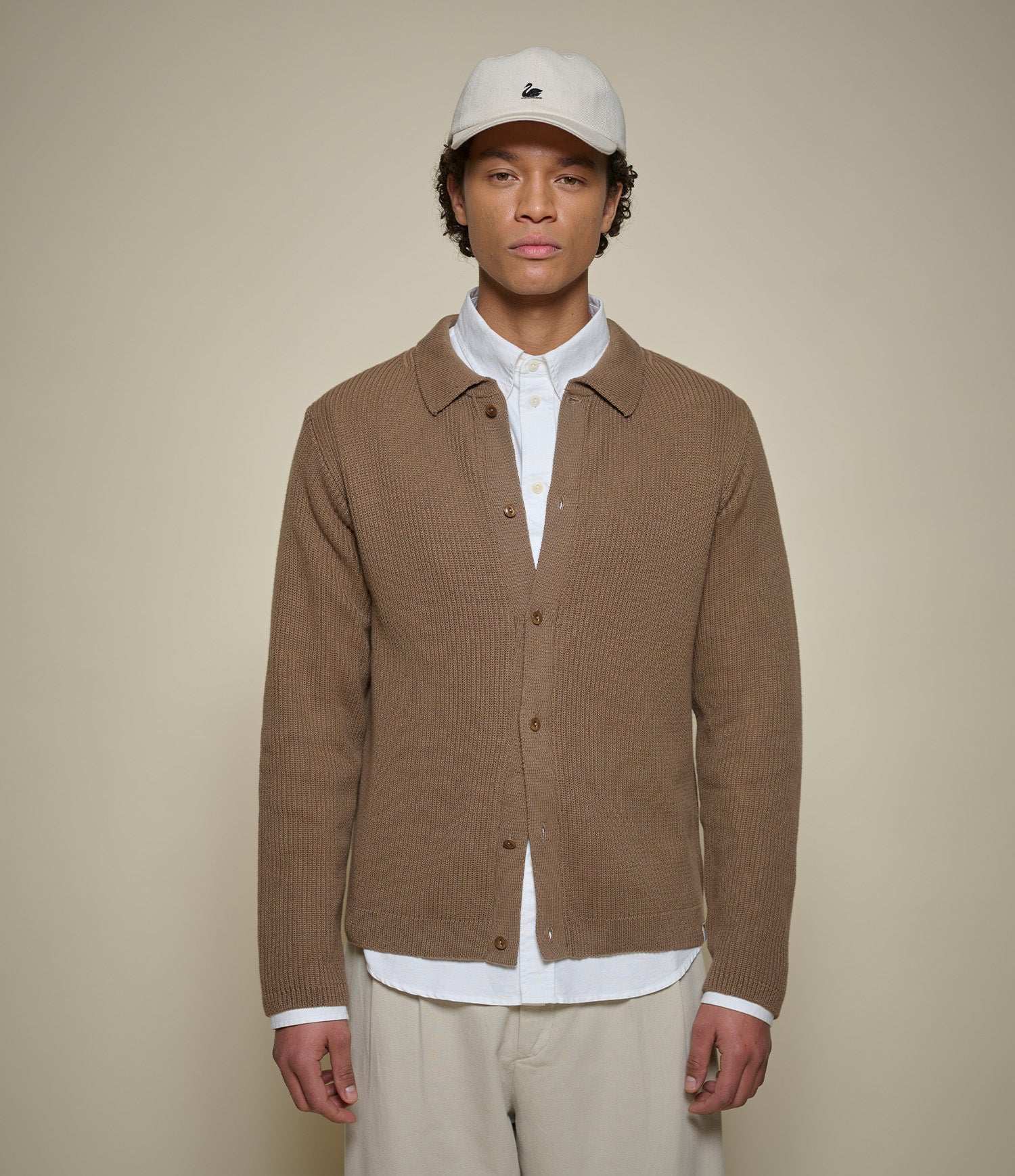 A man wearing a brown ribbed knit button-up cardigan over a white collared shirt, paired with beige trousers and a white baseball cap, standing against a solid beige background.