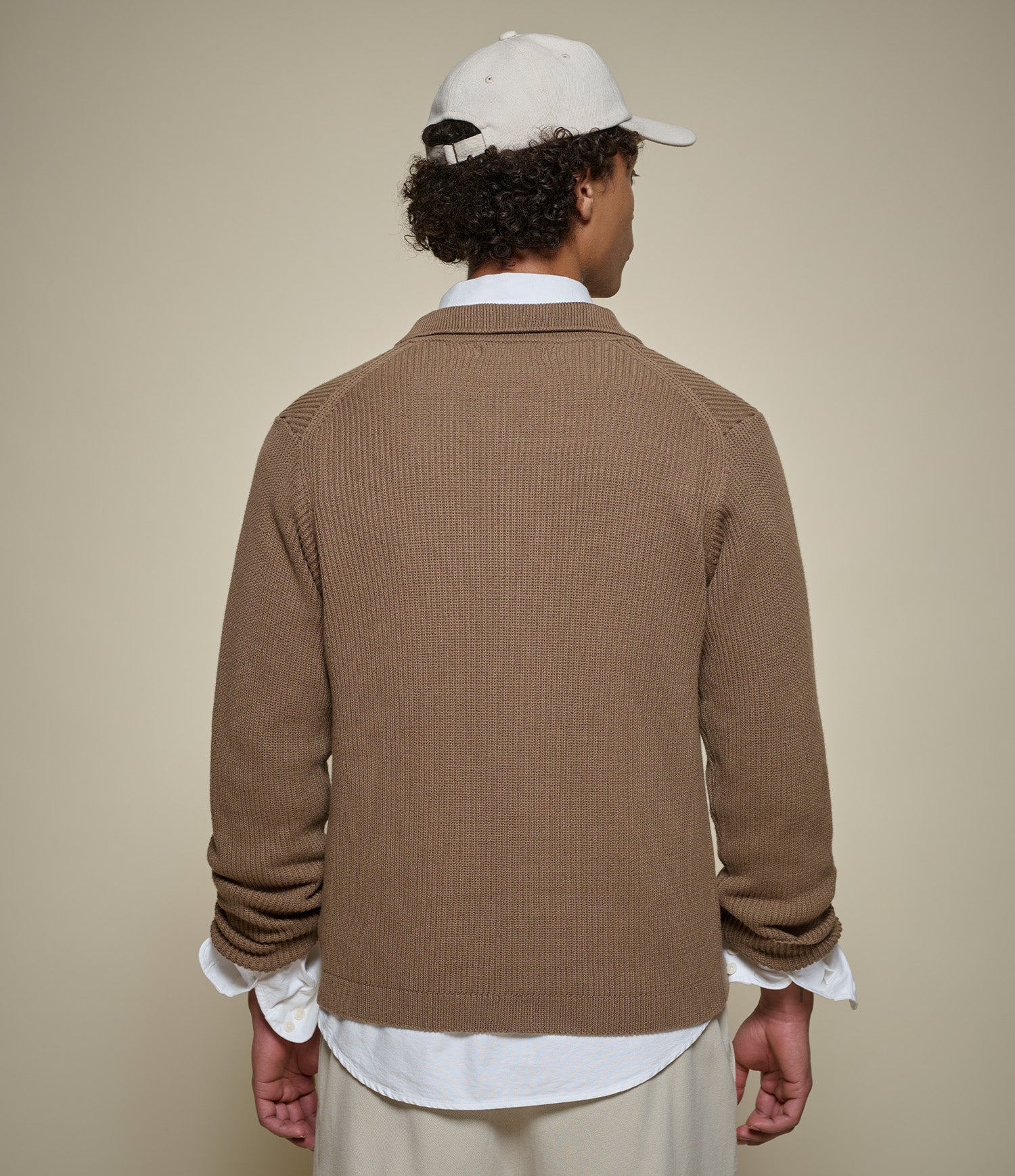 A man viewed from behind wearing a brown ribbed knit cardigan over a white collared shirt, with a white baseball cap and light beige pants, against a plain beige studio background.
