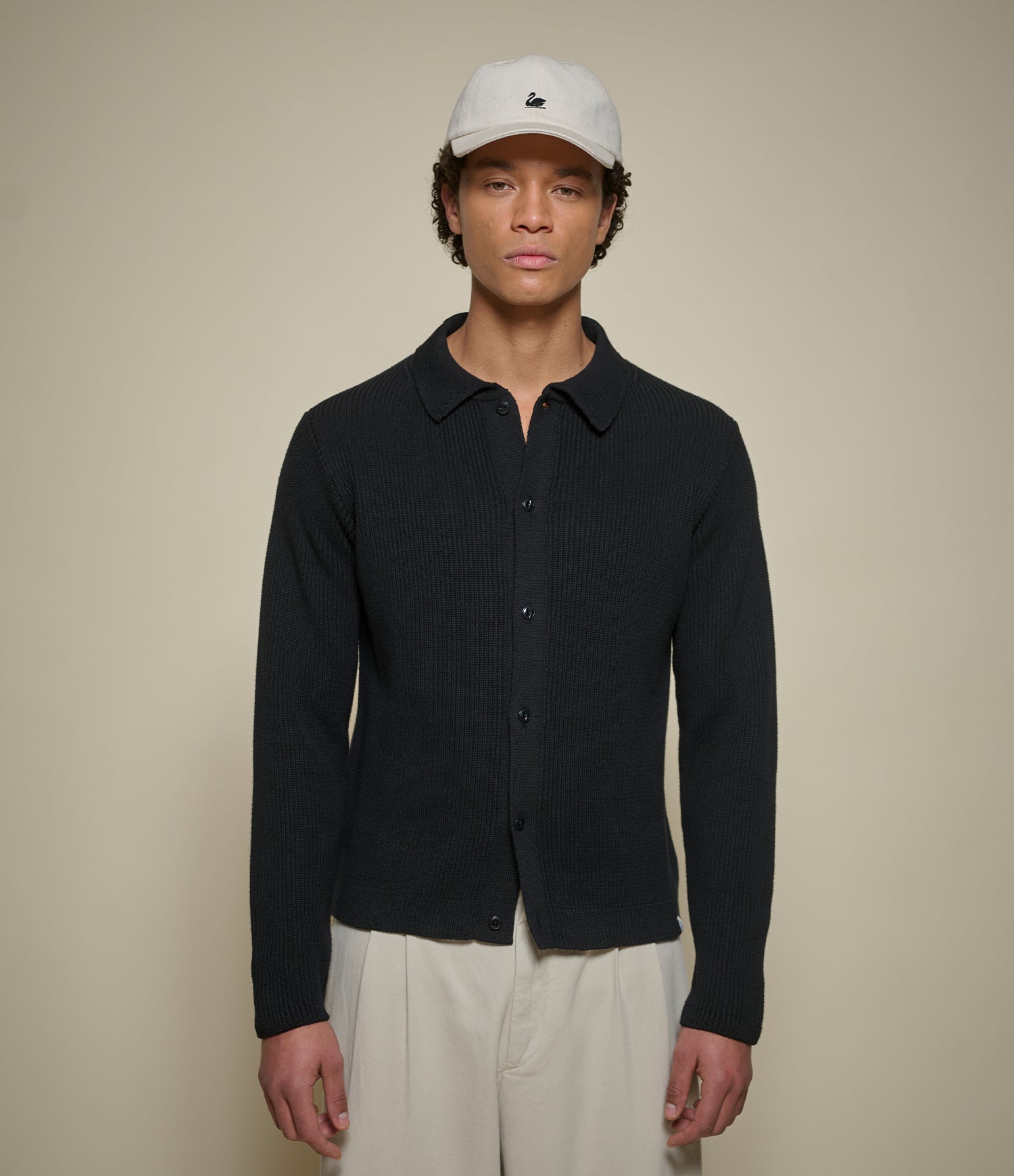 A man wearing a black ribbed knit button-up shirt with a collar, paired with off-white trousers and a white baseball cap, standing against a solid beige background.