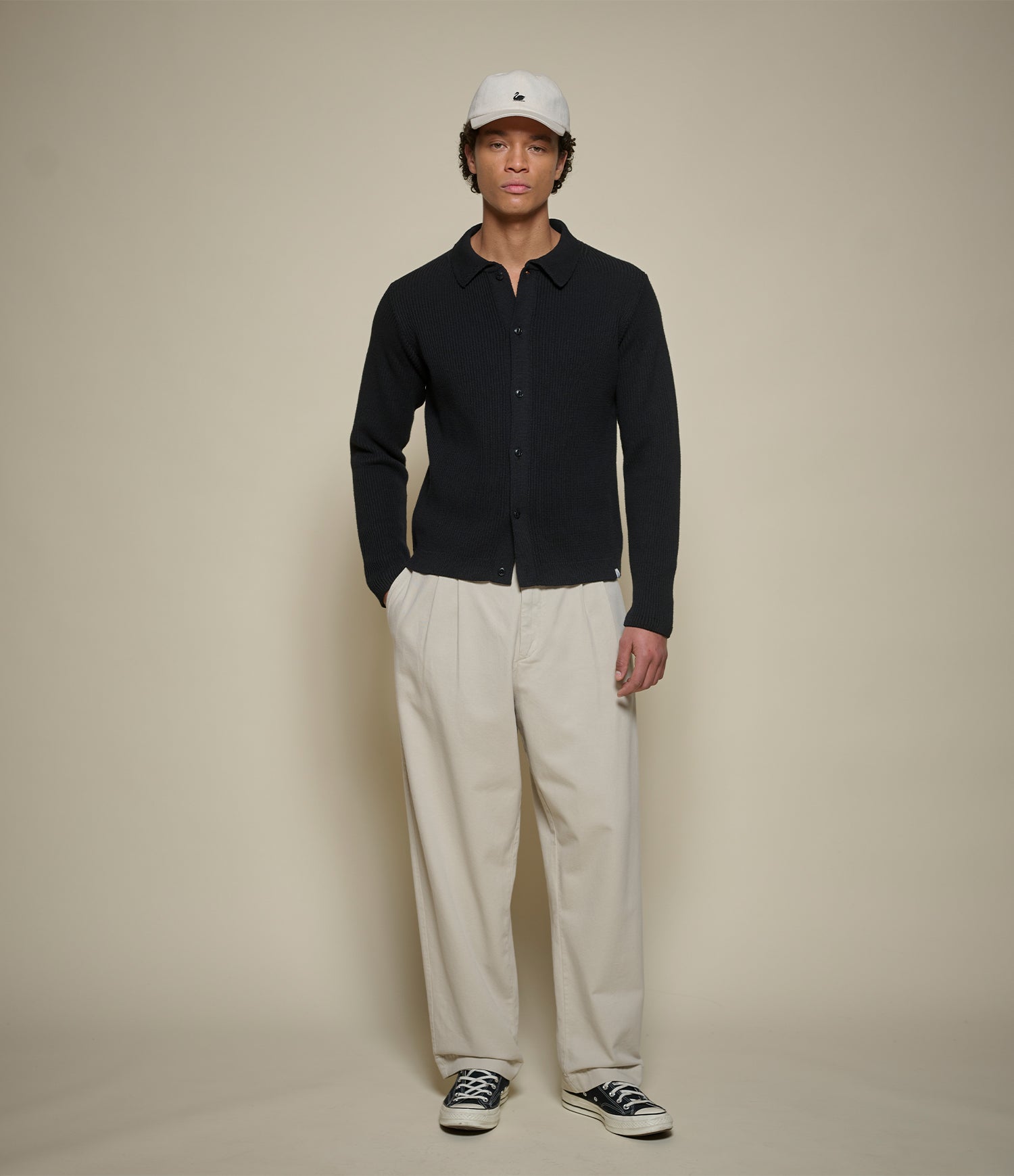 A man stands against a plain beige studio background wearing a black long-sleeve button-up shirt, cream pleated trousers, a white baseball cap, and black-and-white sneakers.