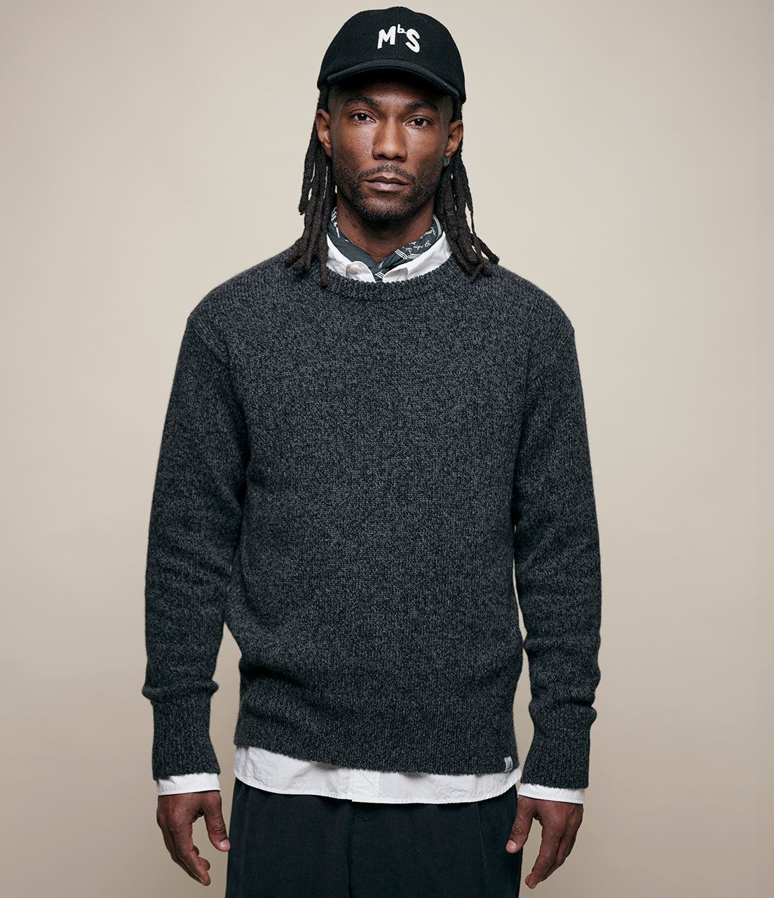 Man wearing a dark gray sweater, white shirt, and black cap with 'MbS' logo on a beige background