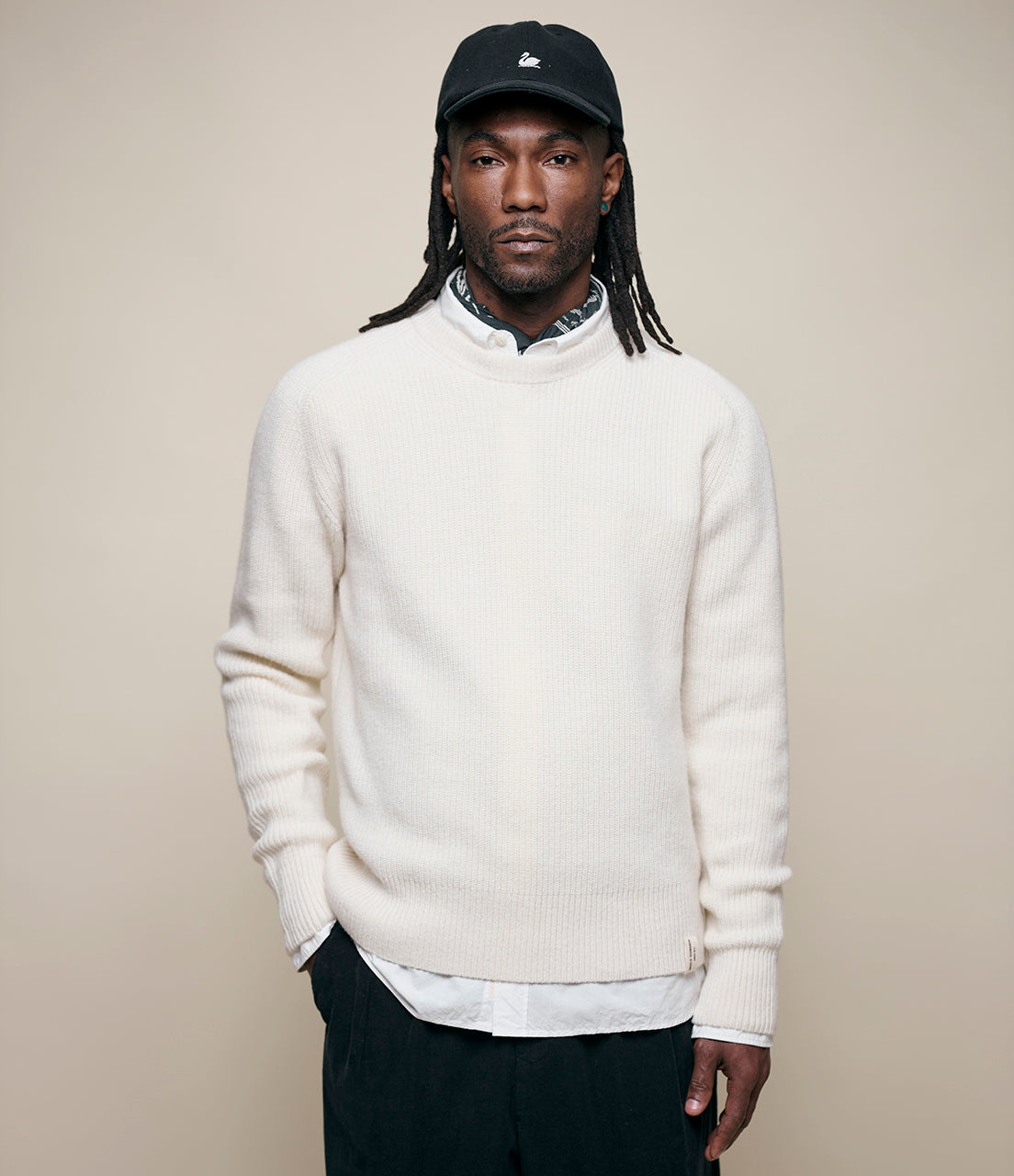Man wearing a white sweater over a collared shirt with a black cap on a beige background