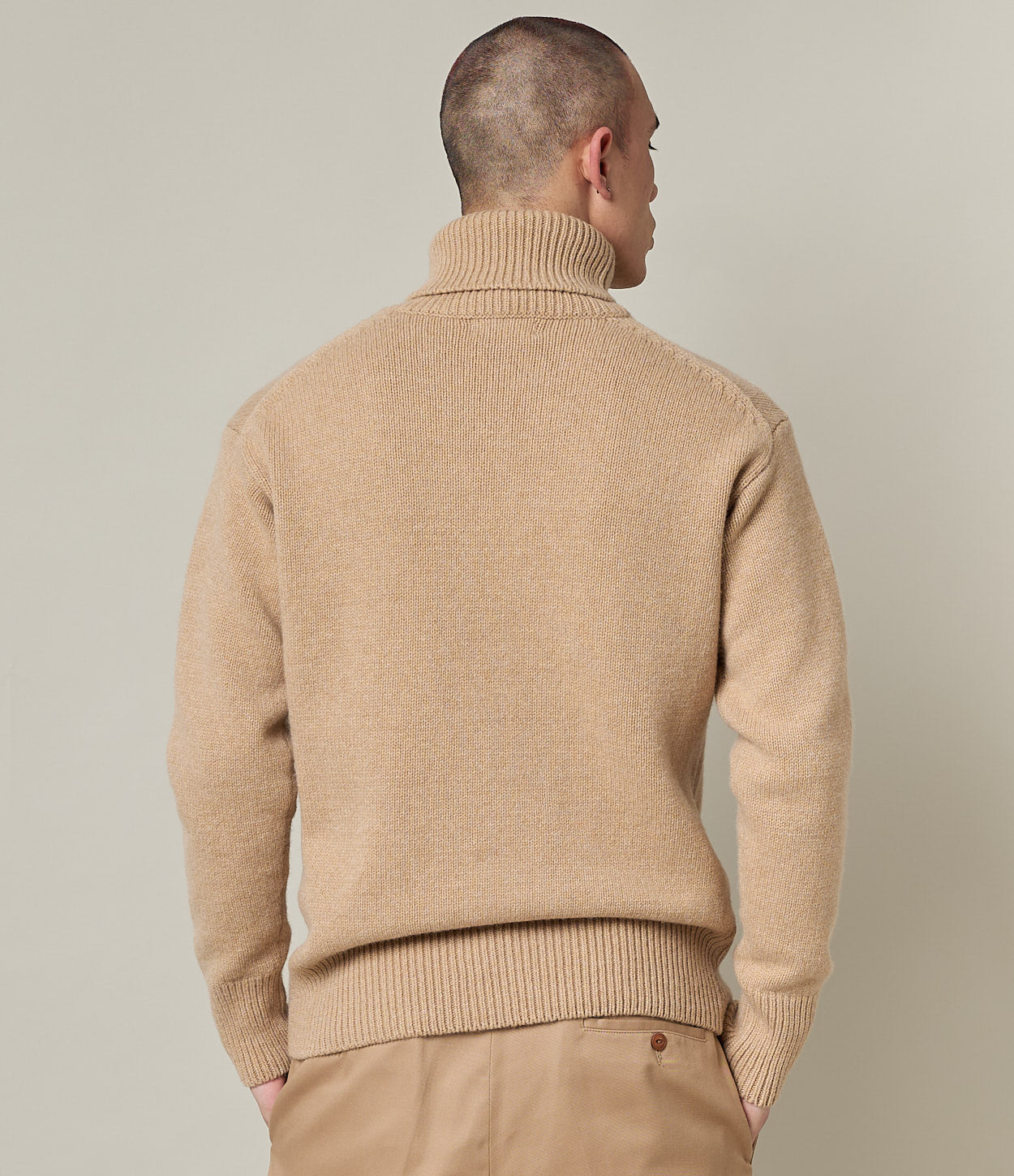 Man wearing a beige turtleneck sweater and pants against a neutral background