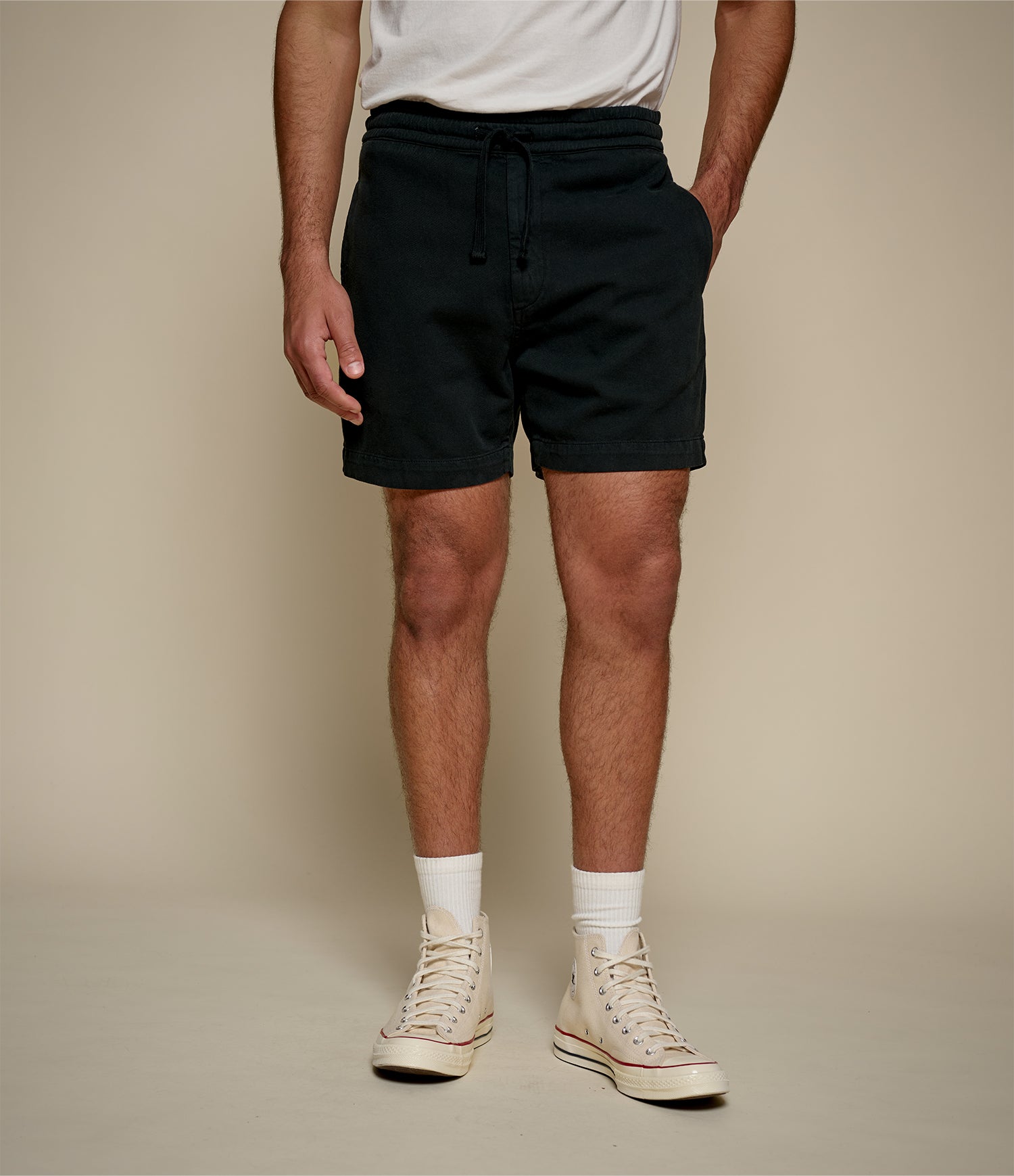 Person wearing black twill shorts, white socks, and beige high-top sneakers on a plain background