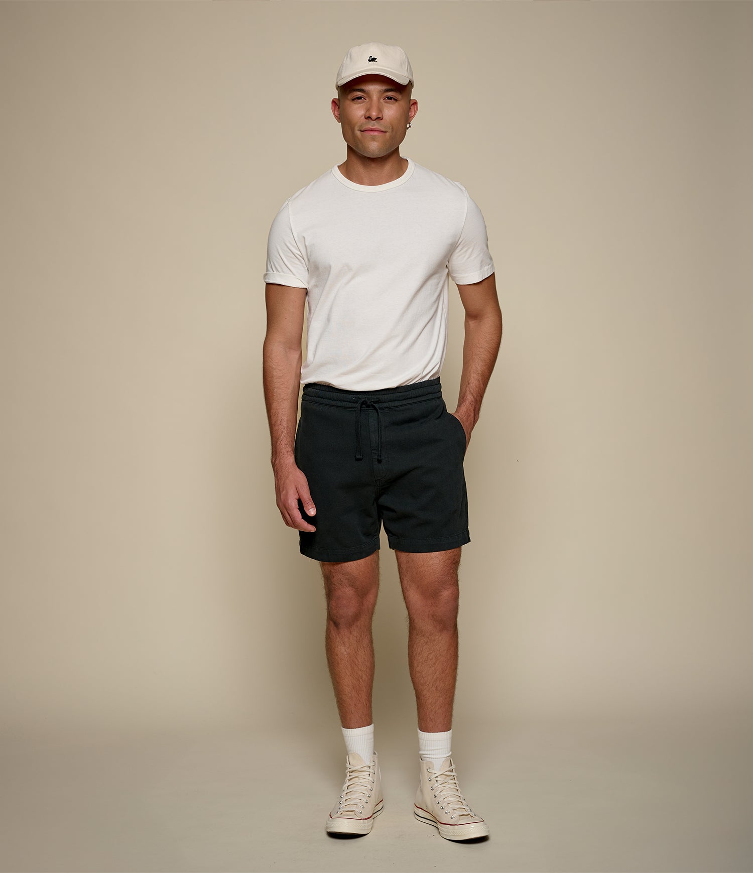 Man wearing a white t-shirt, black twill shorts, and a beige cap on a beige background