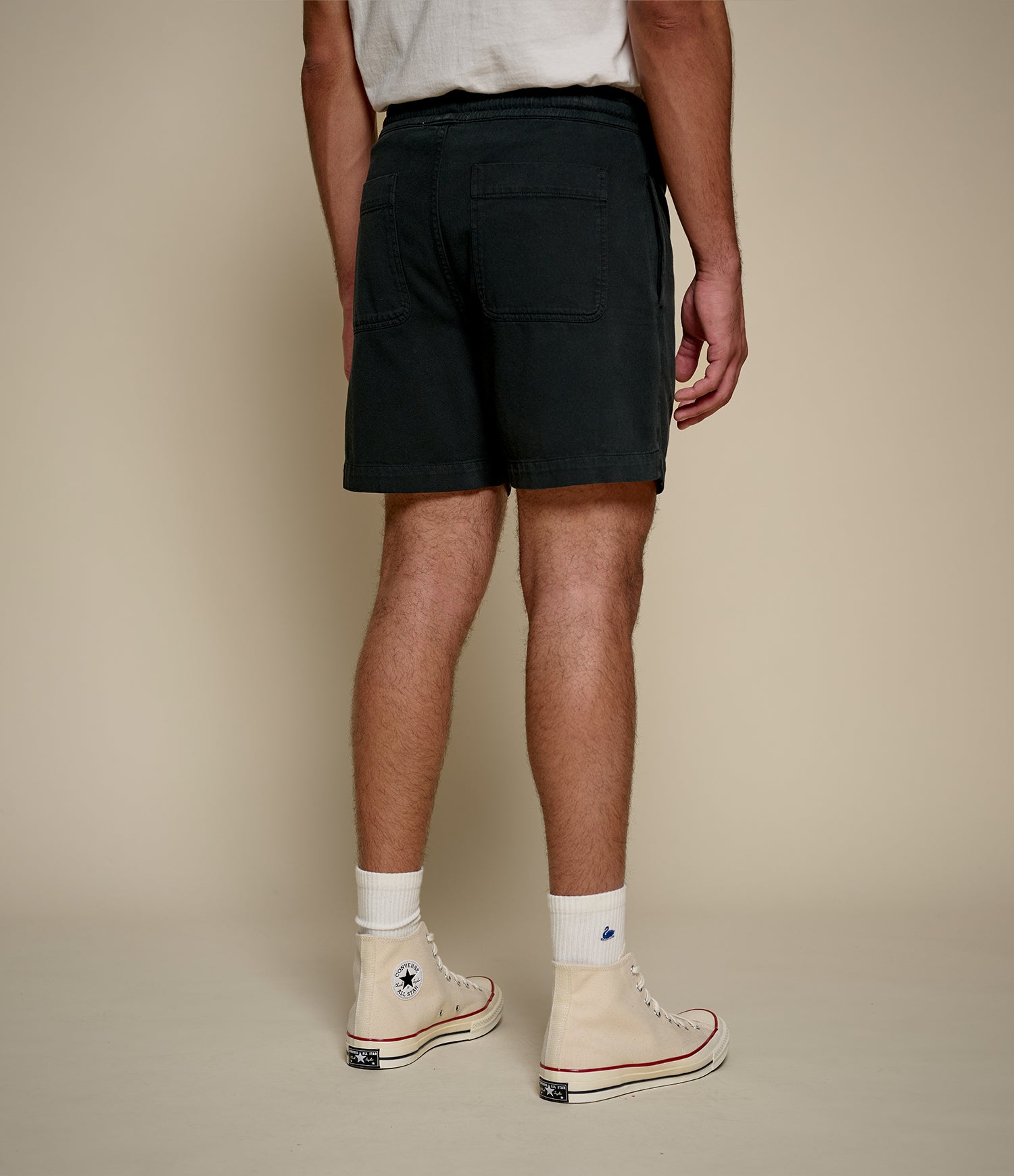 Person wearing black twill shorts and beige Converse shoes on a plain background