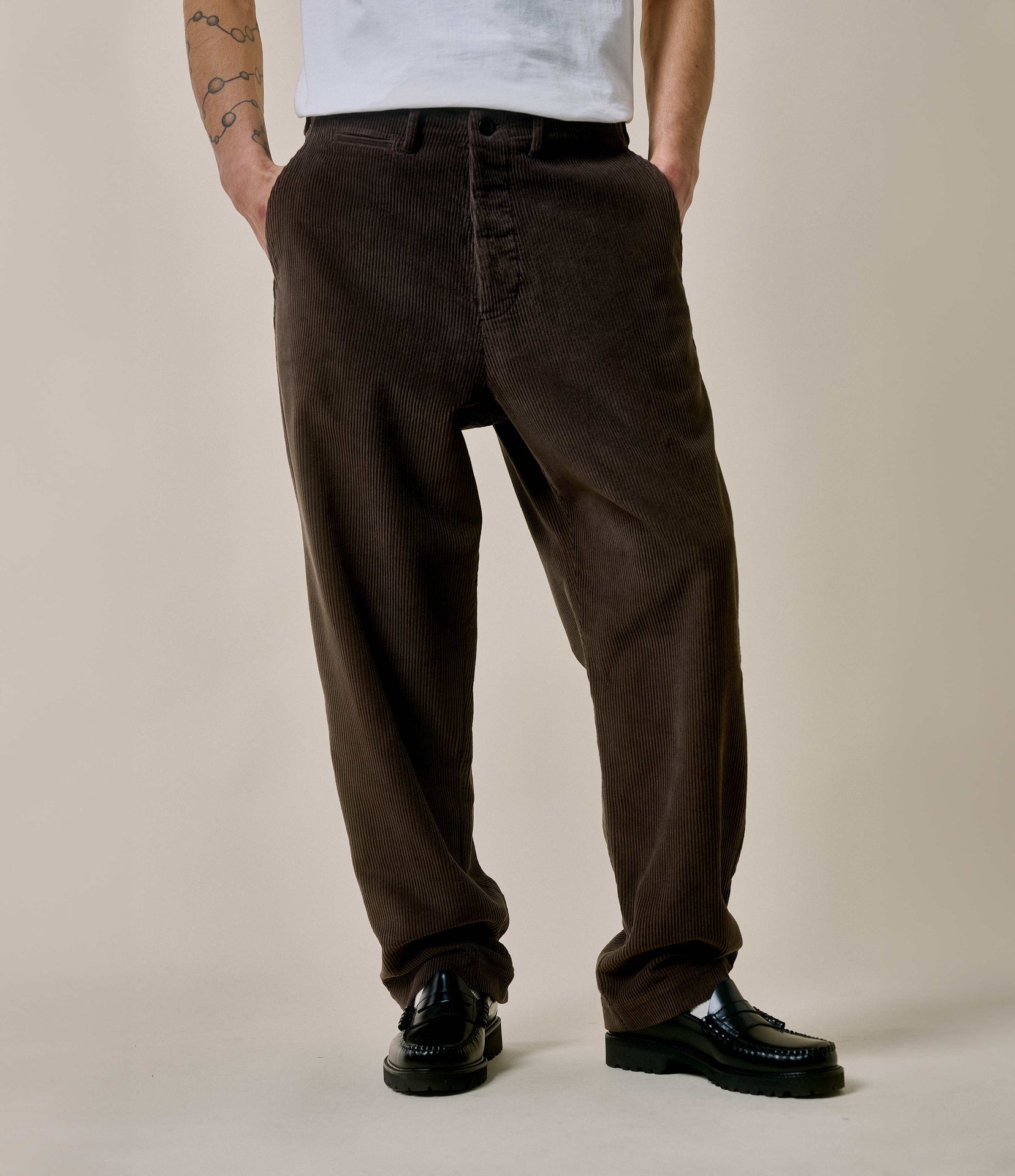 A person standing against a plain background, wearing brown corduroy chino pants with a relaxed fit and pleats, paired with a white T-shirt and black shoes.
