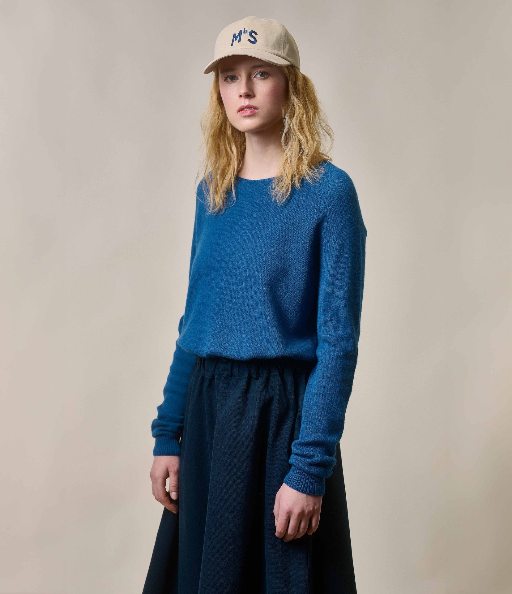 A person wearing a blue long-sleeve pullover with a widened round-neck, paired with a dark skirt and a beige cap with a logo in the front.
