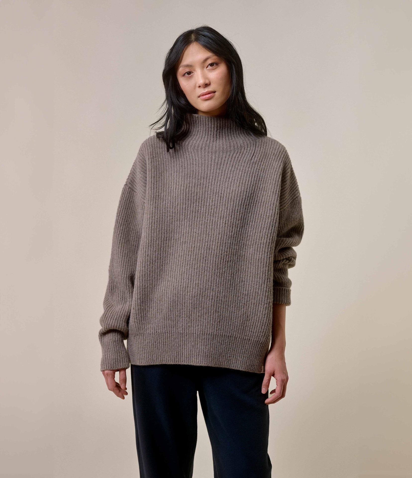 A person wearing a brown chunky ribbed merino wool-cashmere mock-neck sweater with black pants.