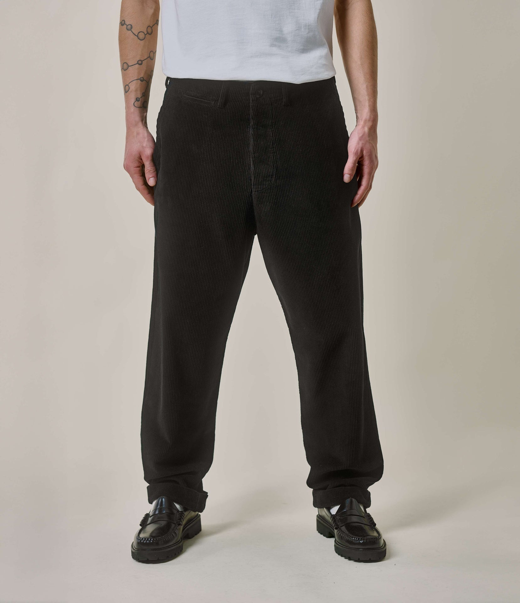 A person standing against a plain background, wearing black corduroy chino pants with a relaxed fit and pleats, paired with a white T-shirt and black shoes.