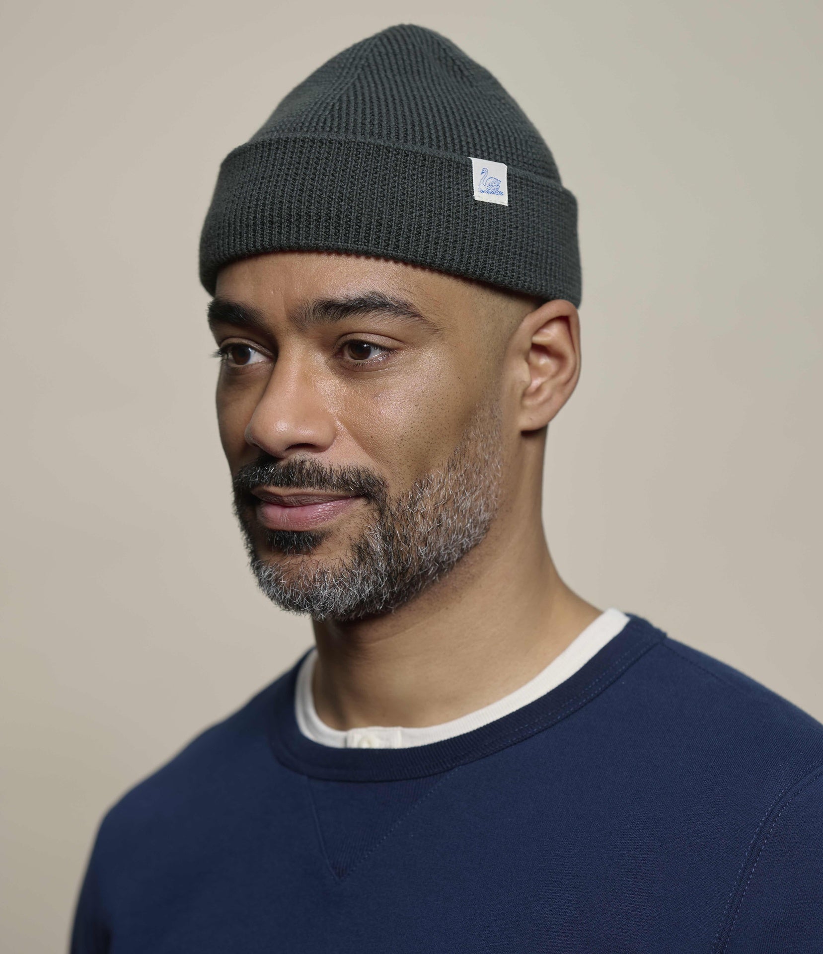 A man wearing a green ribbed knit beanie with a small white square logo patch on the cuff, paired with a navy blue crewneck sweatshirt. The background is a solid, warm beige studio backdrop.