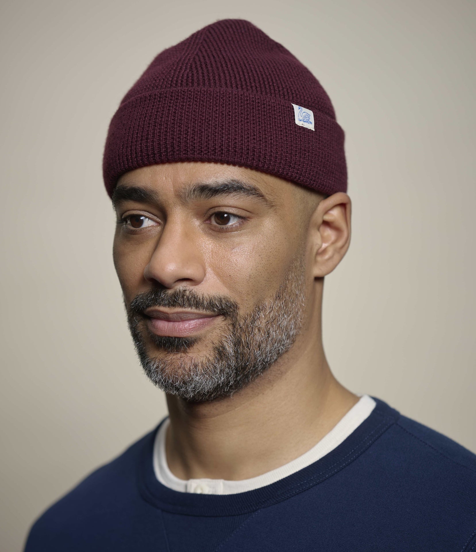 A man wearing a burgundy knit beanie with a small white rectangular logo patch on the side, photographed against a plain light beige background.