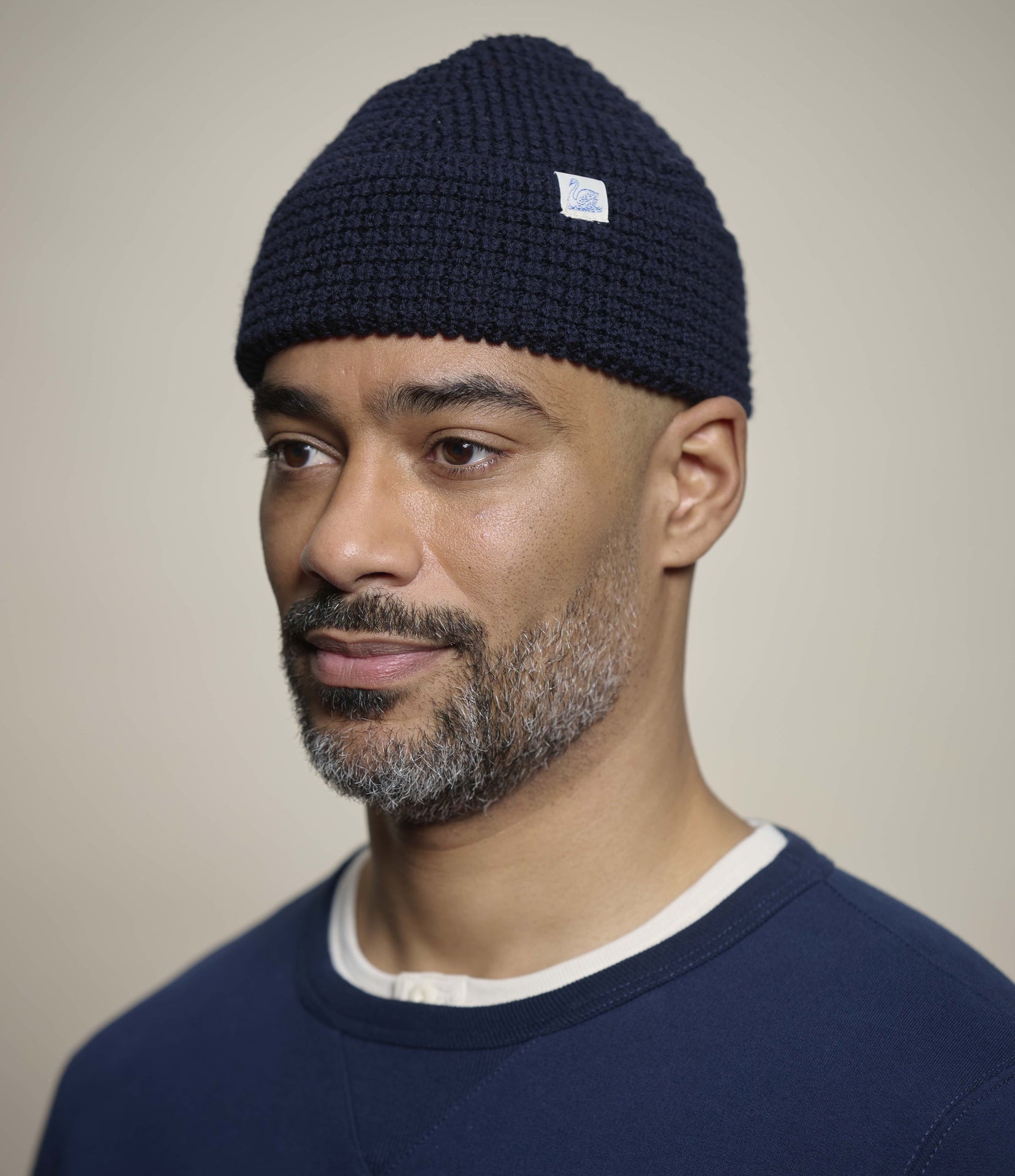 A man wearing a dark navy blue knit beanie with a small white square logo patch on the side, paired with a navy crewneck sweater against a plain beige background.