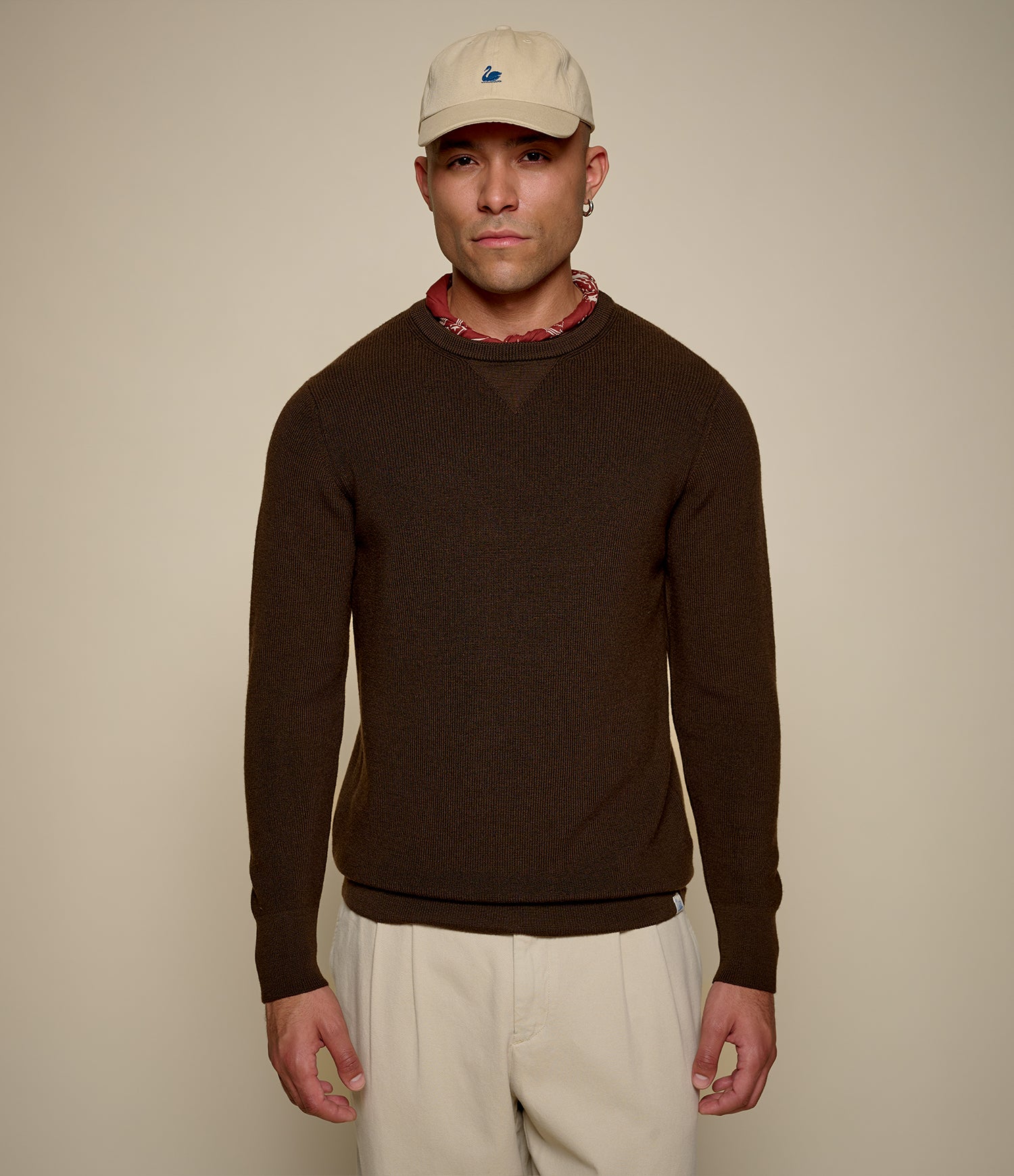 A man wearing a dark brown long-sleeve knit sweater, cream pants, a beige baseball cap, and a red patterned neck gaiter, standing against a solid beige background.