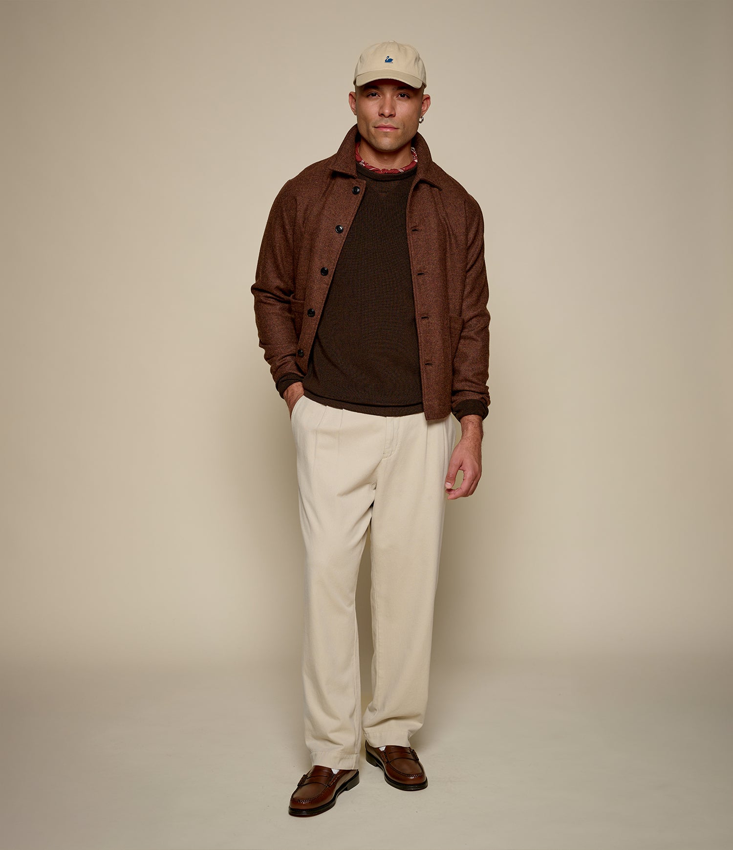 A man modeling a brown tweed jacket over a dark brown shirt, cream trousers, and brown loafers, wearing a beige baseball cap against a solid beige studio background.
