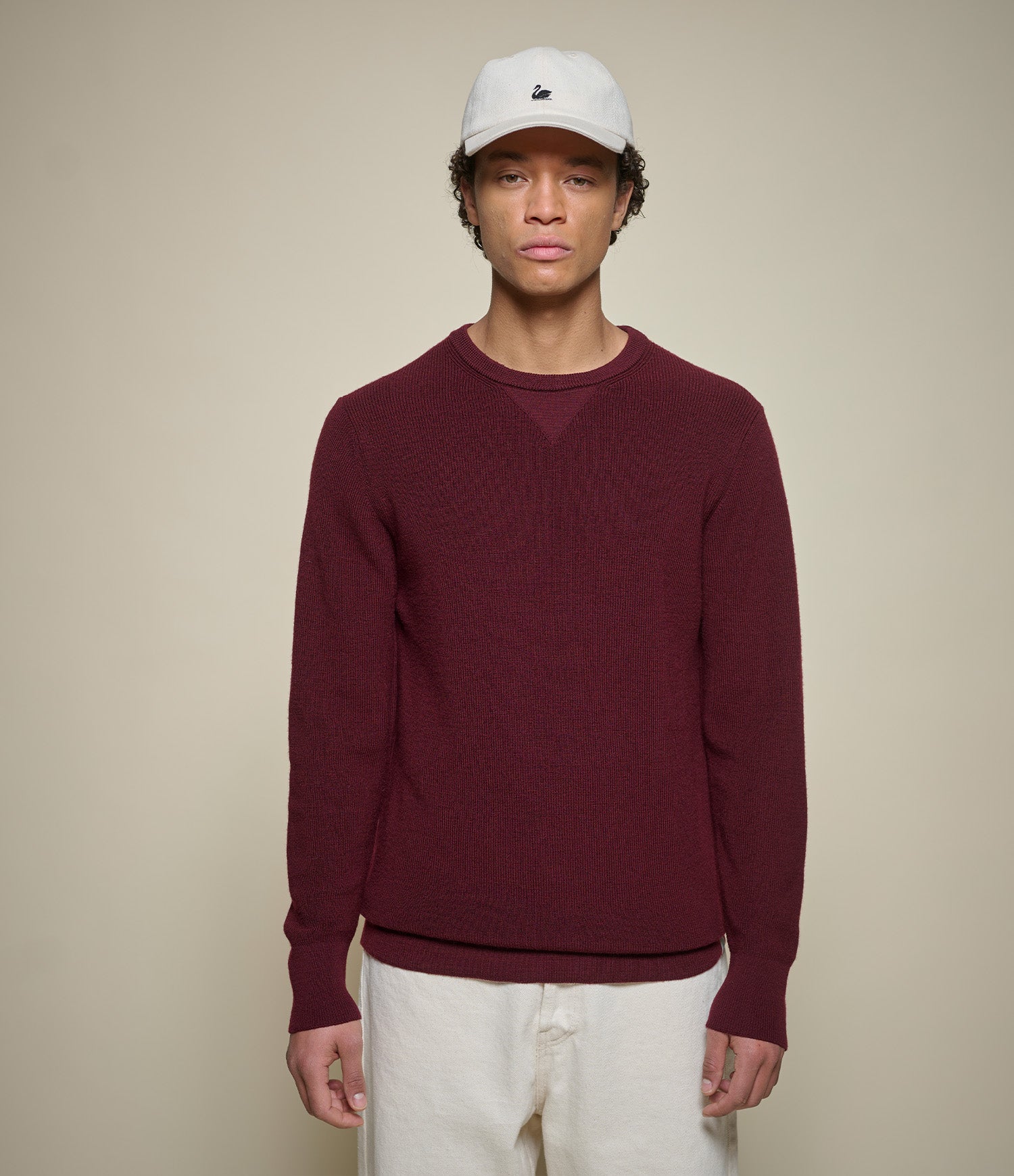 Person wearing a burgundy merino wool sweater and white cap against a beige background