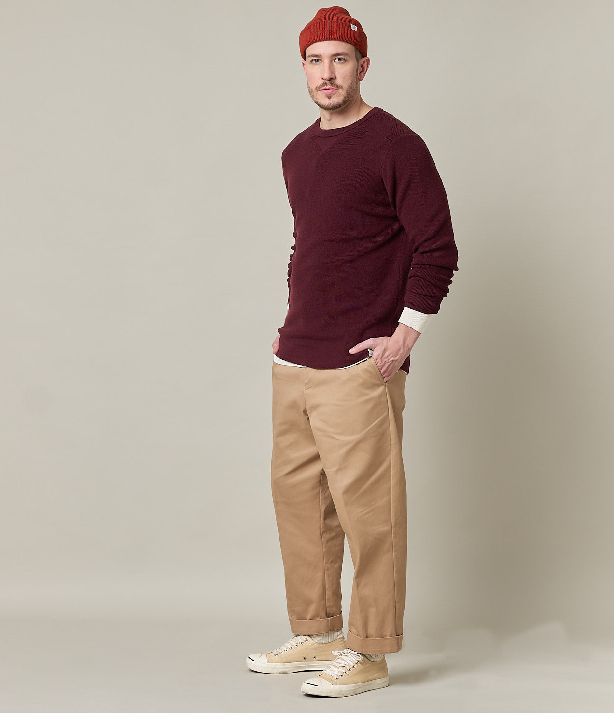 Man wearing a burgundy sweater, beige pants, and a red beanie against a plain background