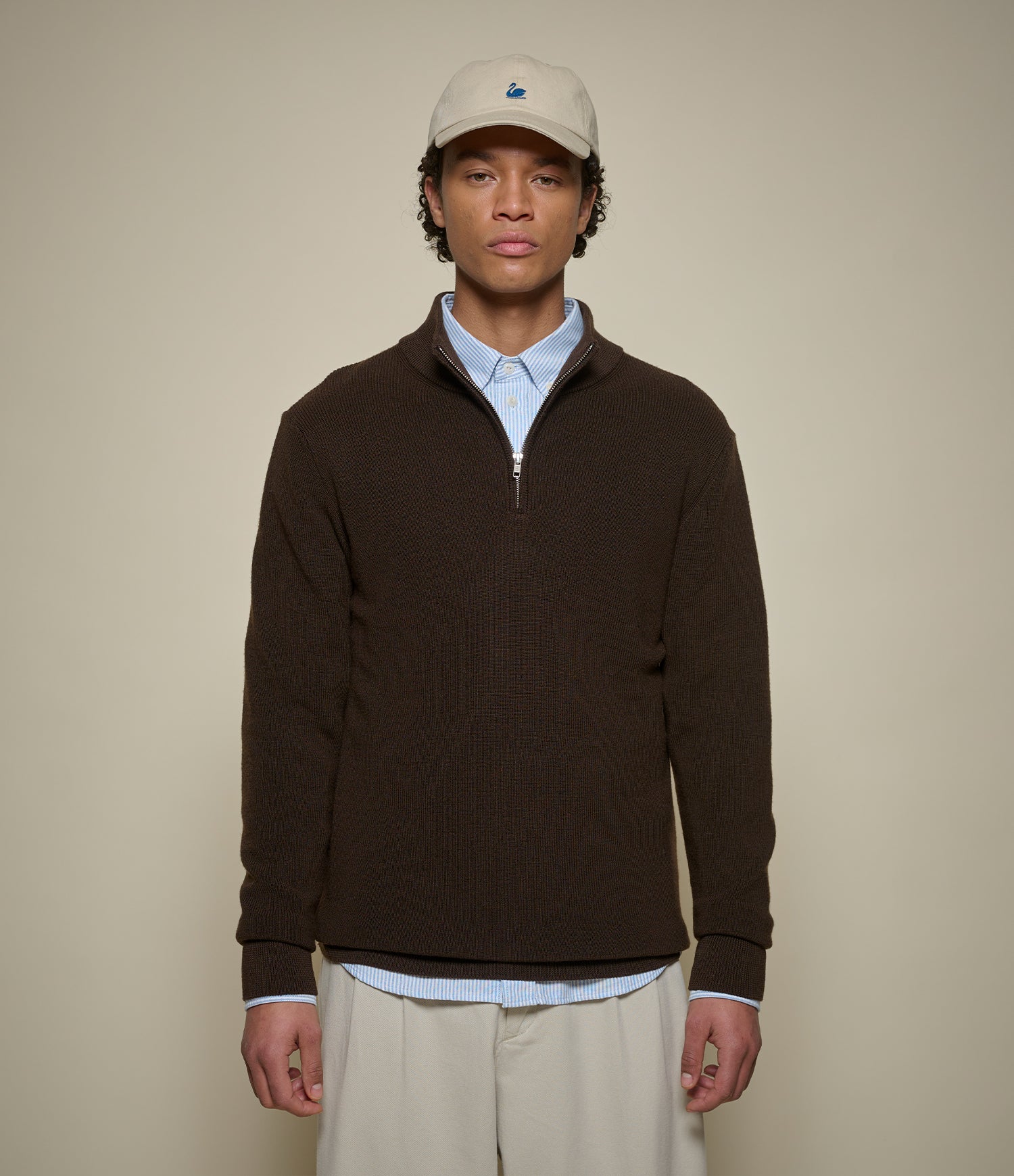 Man wearing a brown half-zip sweater, light blue shirt, beige pants, and a beige cap with a logo on a beige background