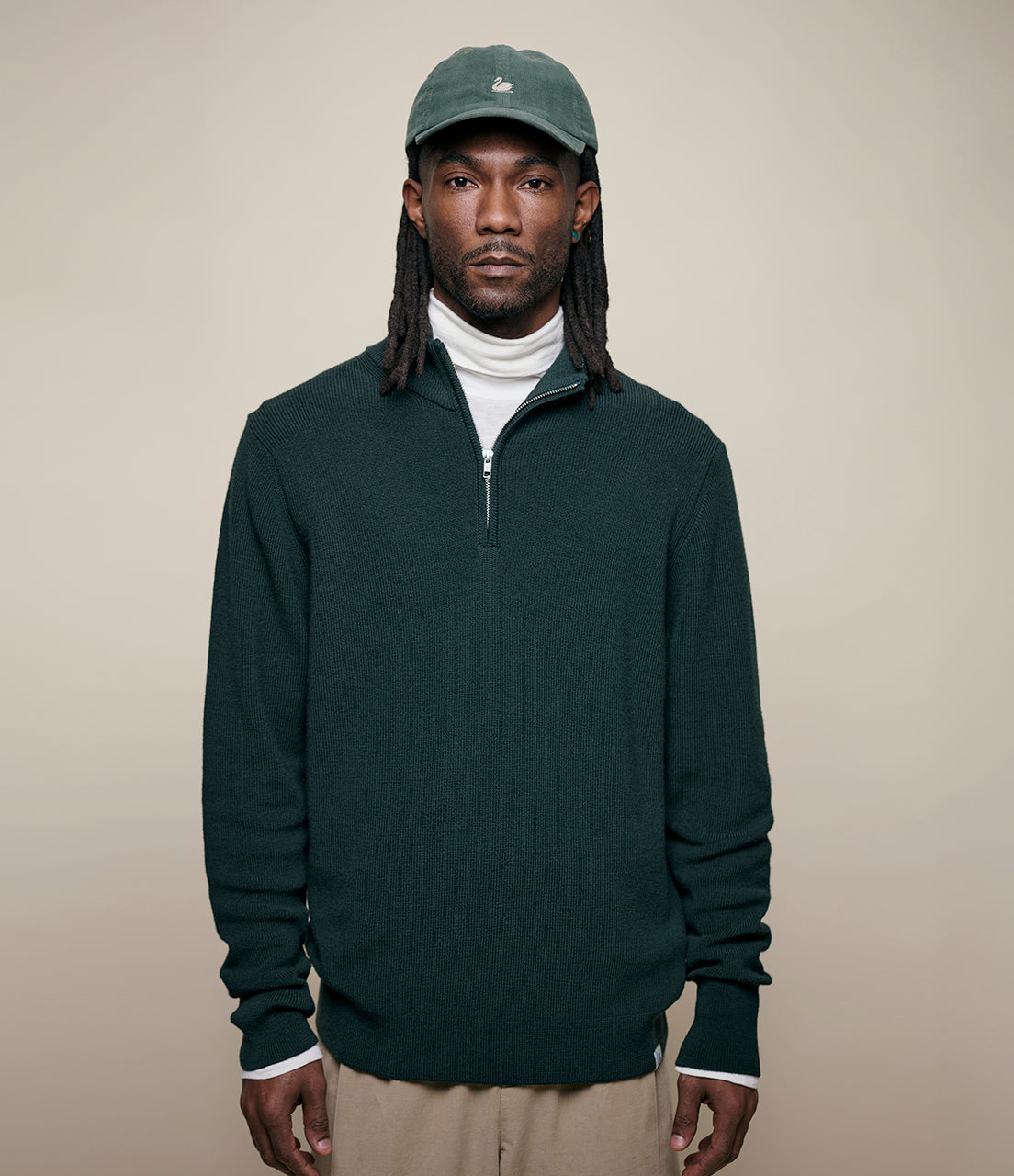 Man wearing a dark green half-zip sweater and cap against a beige background