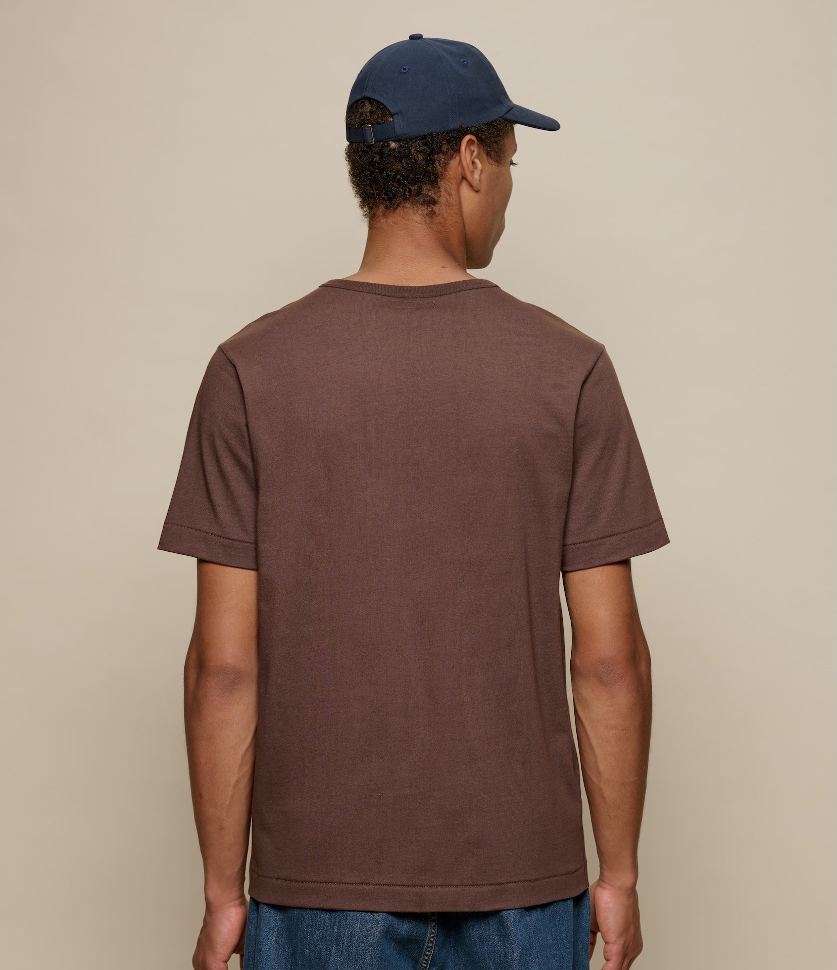 Person wearing a brown t-shirt and blue cap from behind on a beige background