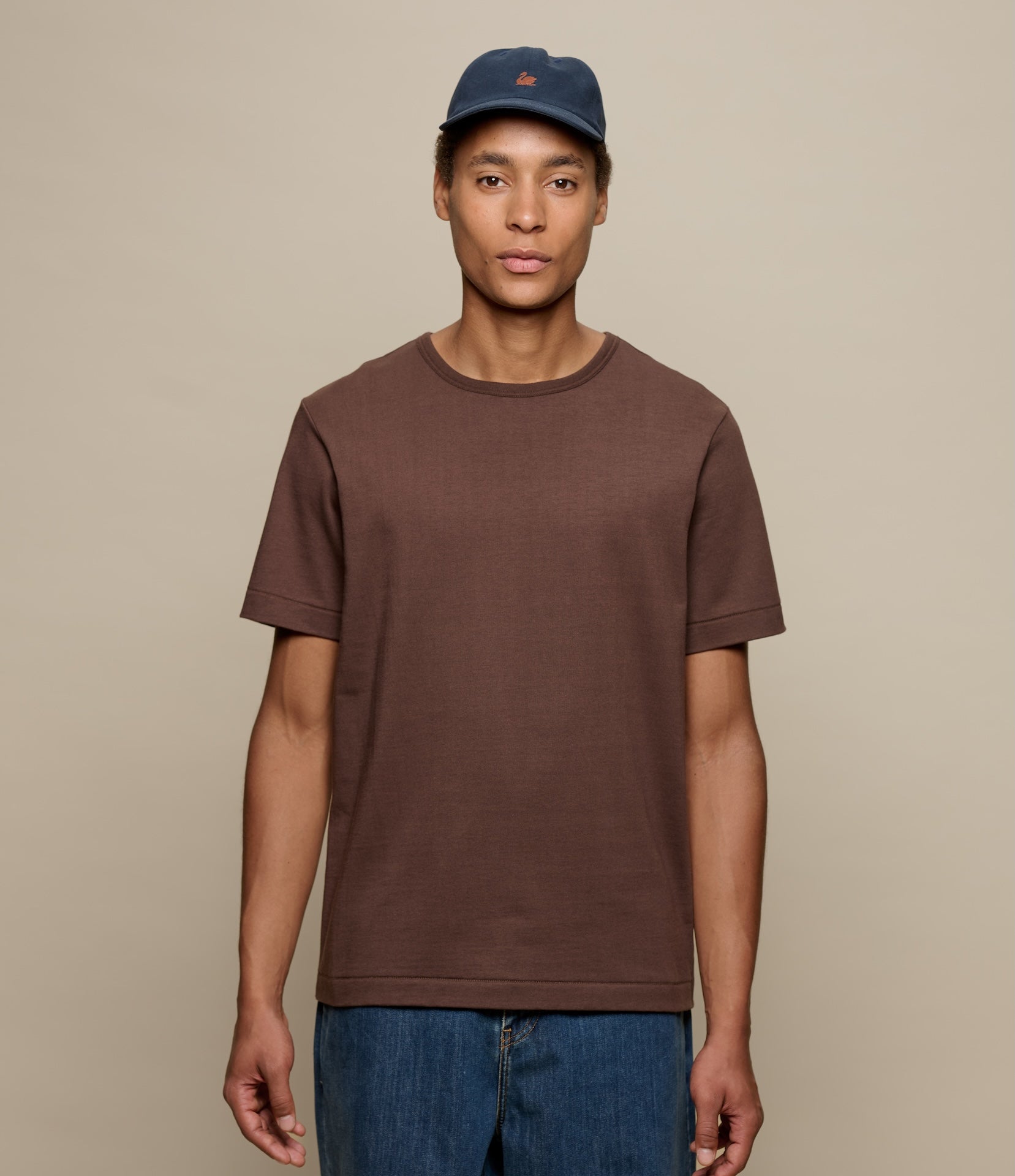 Man wearing a brown t-shirt and blue cap against a beige background