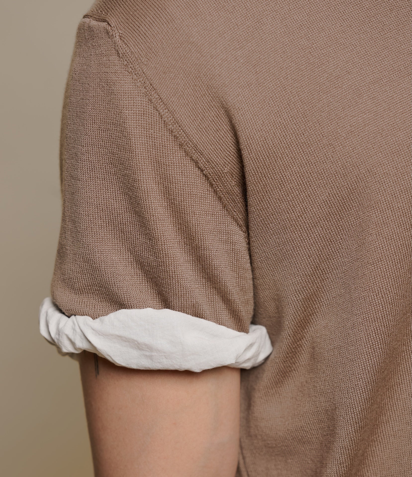 Close-up of a person wearing a brown knitted polo shirt with rolled-up white sleeves against a beige background