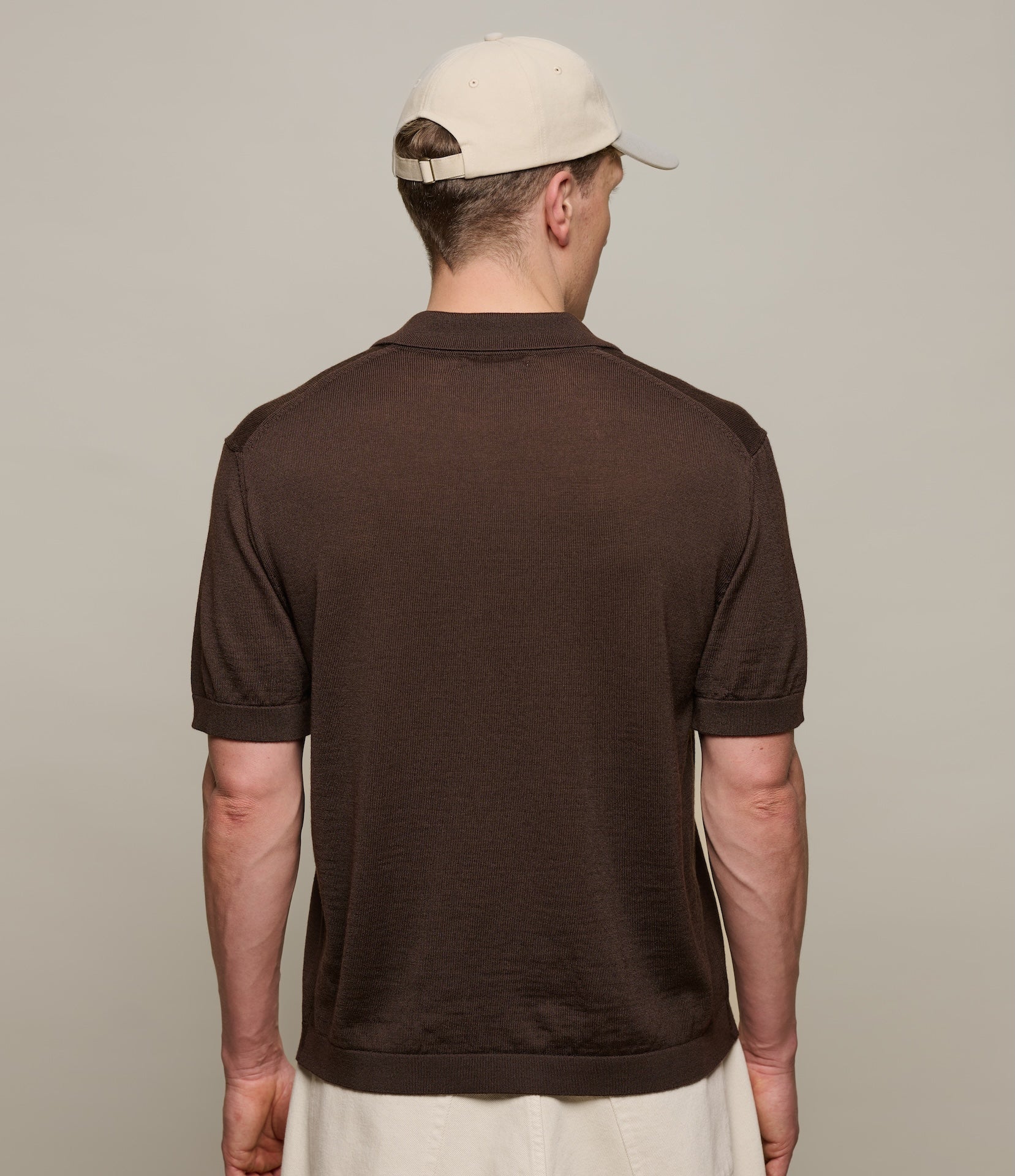 Man wearing a brown knitted polo shirt and beige cap from behind on a plain background