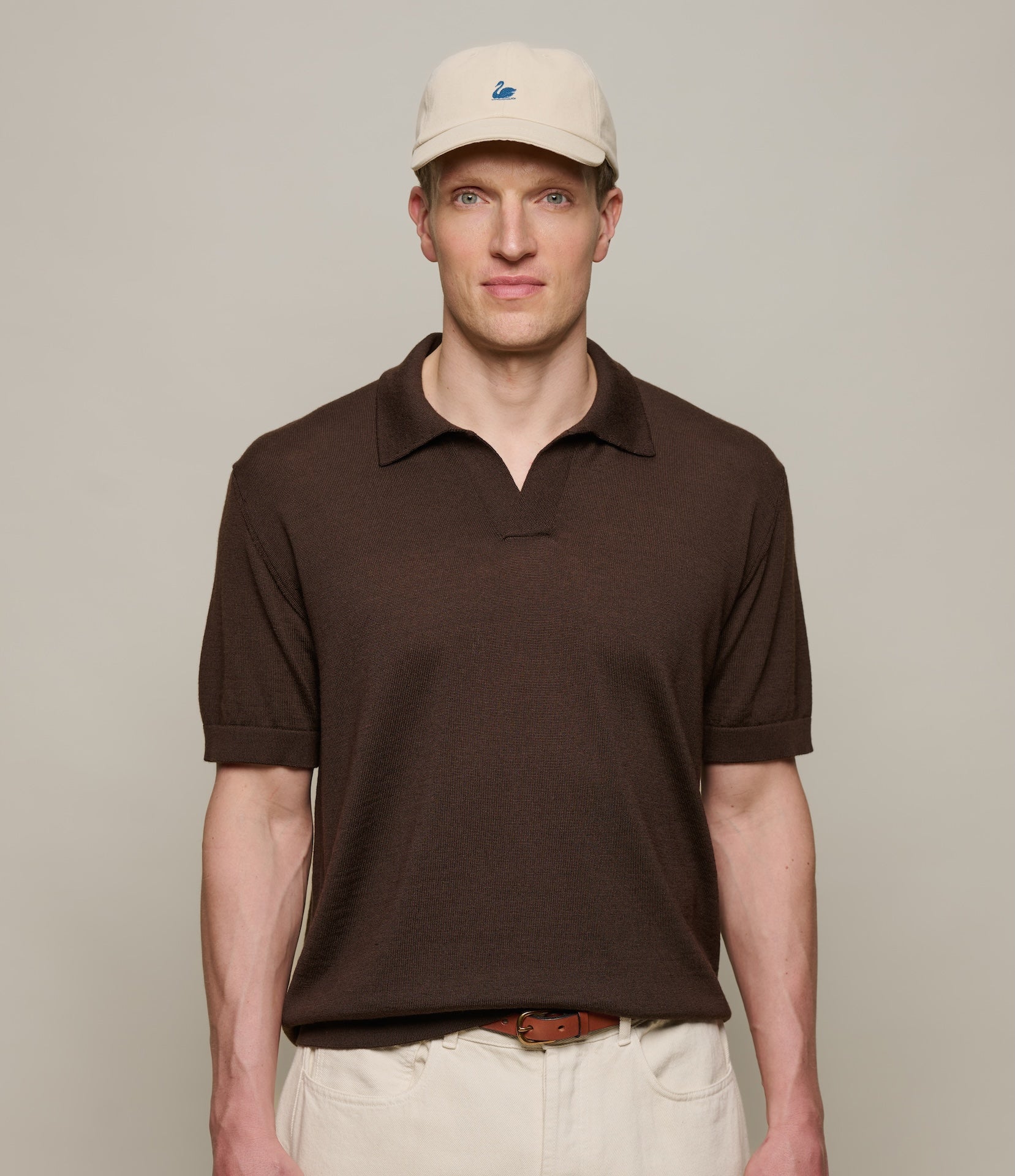 Man wearing a brown knitted merino wool polo shirt and beige cap against a plain background