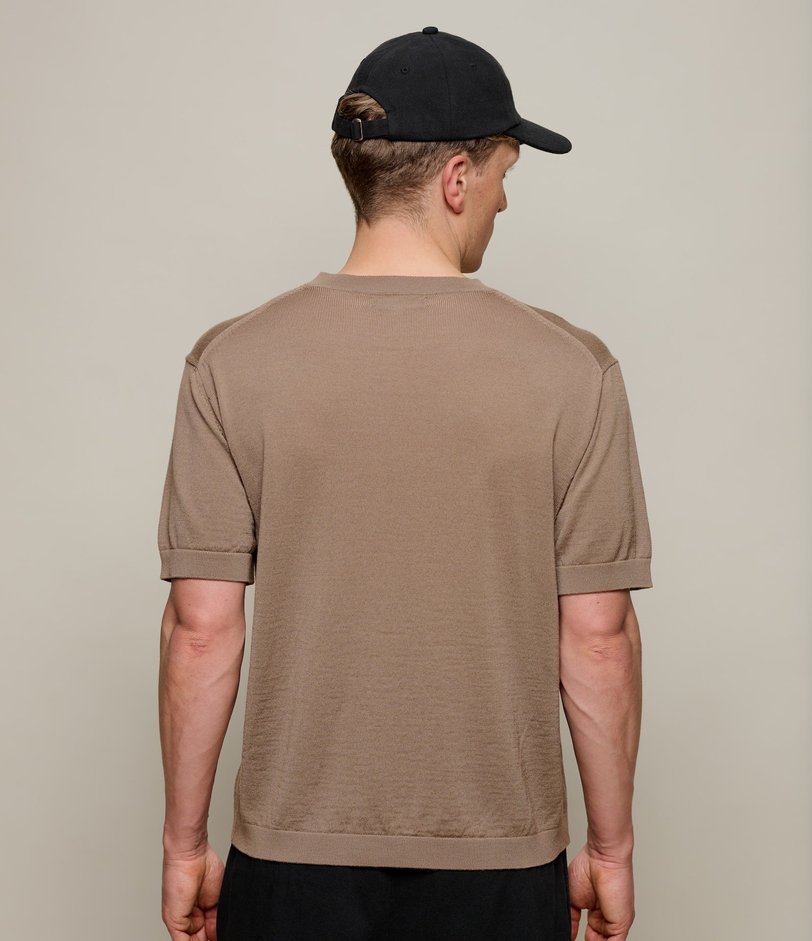 Man wearing a brown knitted t-shirt and black cap from behind on a plain background