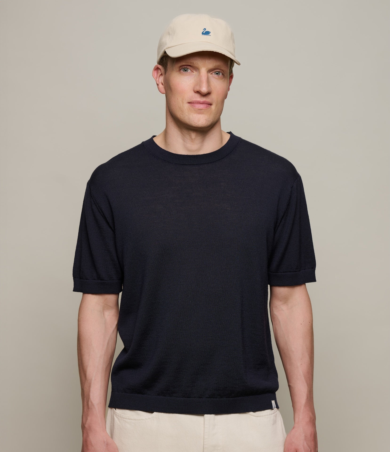 Man wearing a dark navy knitted t-shirt and beige cap against a plain background