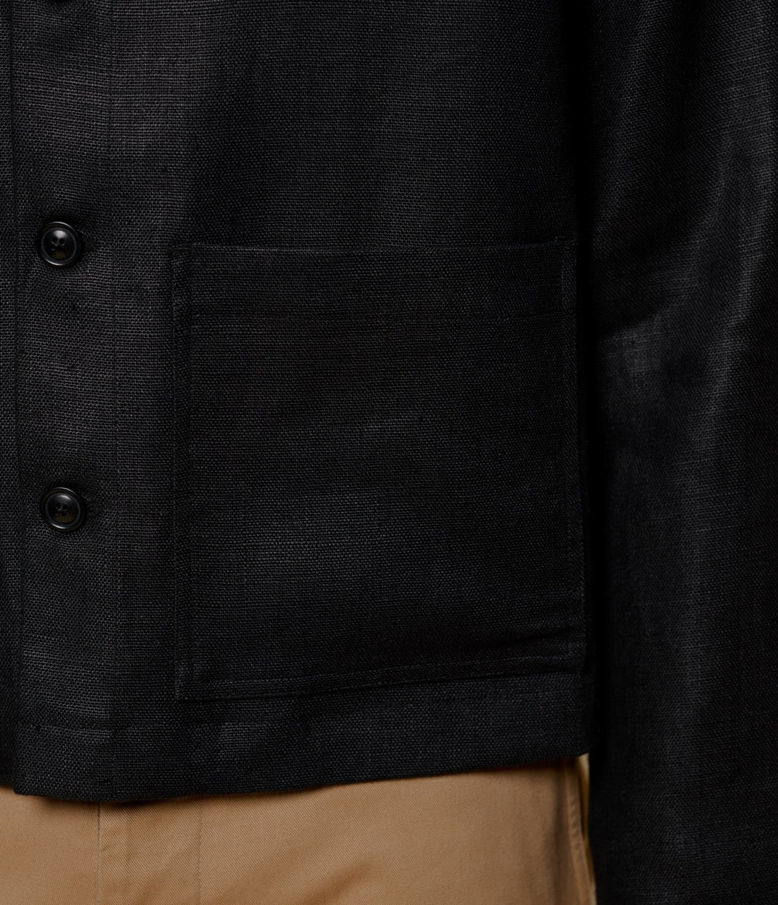 Close-up of a black linen jacket with buttons and a pocket, worn over beige pants.