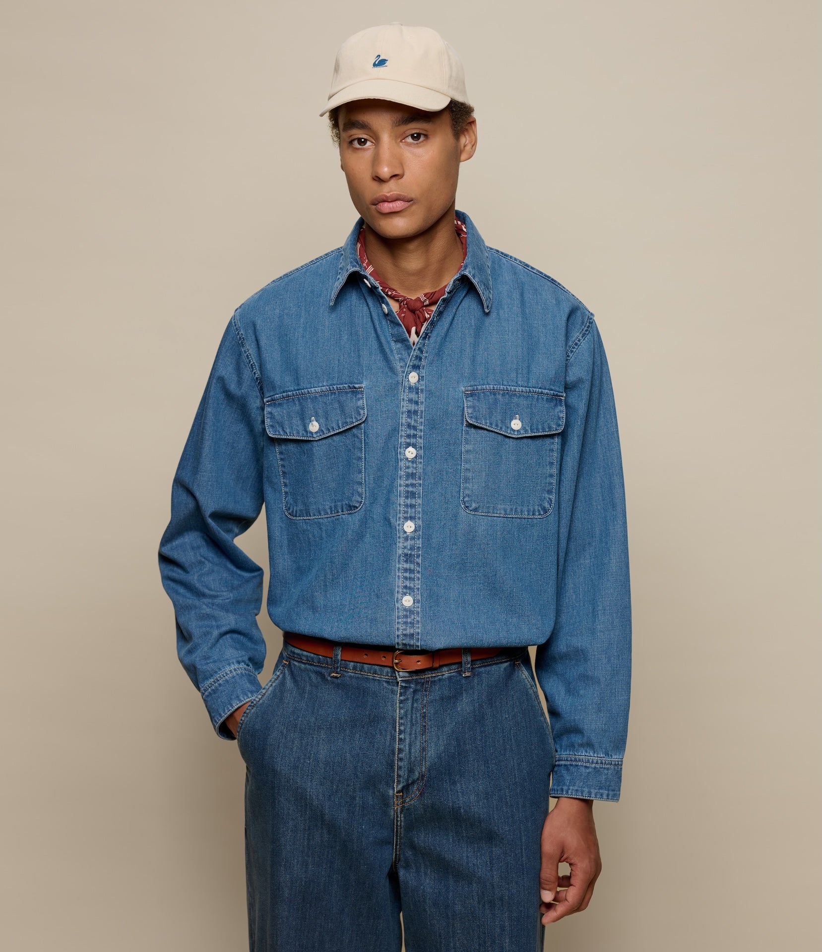 Man wearing a blue denim shirt and pants with a beige cap on a beige background