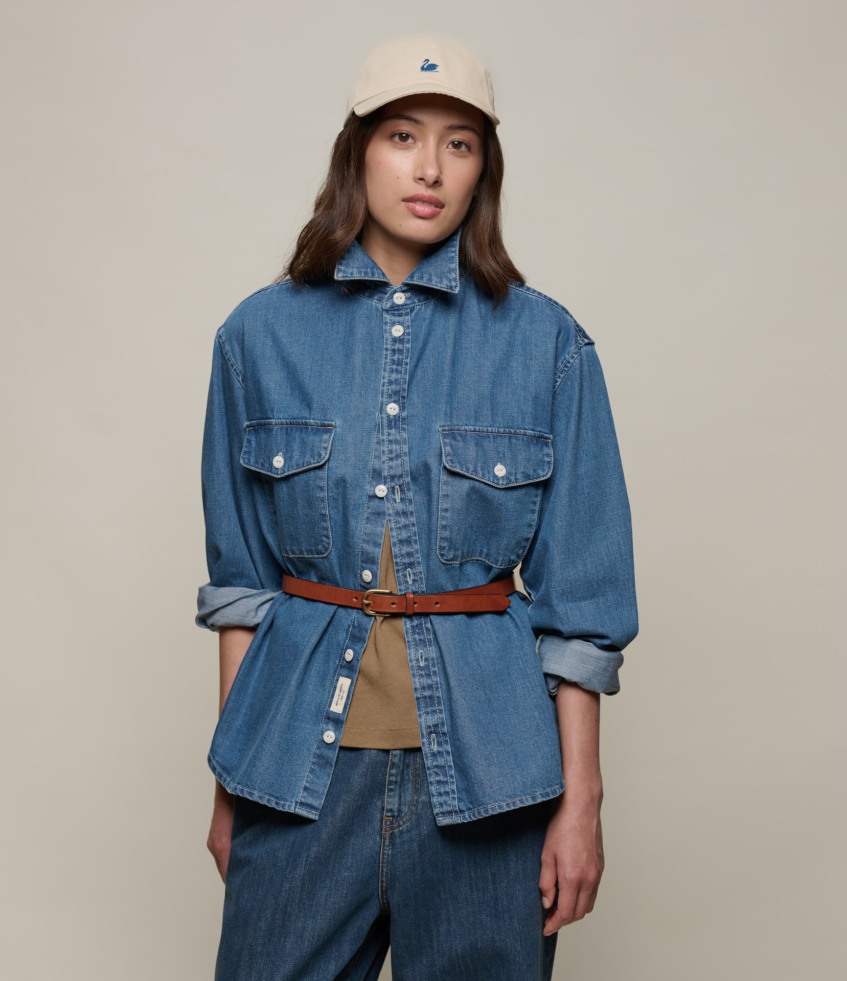 Person wearing a blue denim shirt and jeans with a beige cap on a plain background