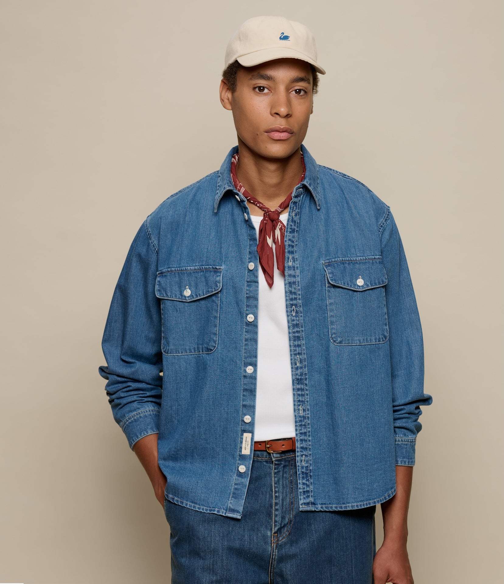 Person wearing a blue denim shirt, beige cap, and red scarf against a beige background