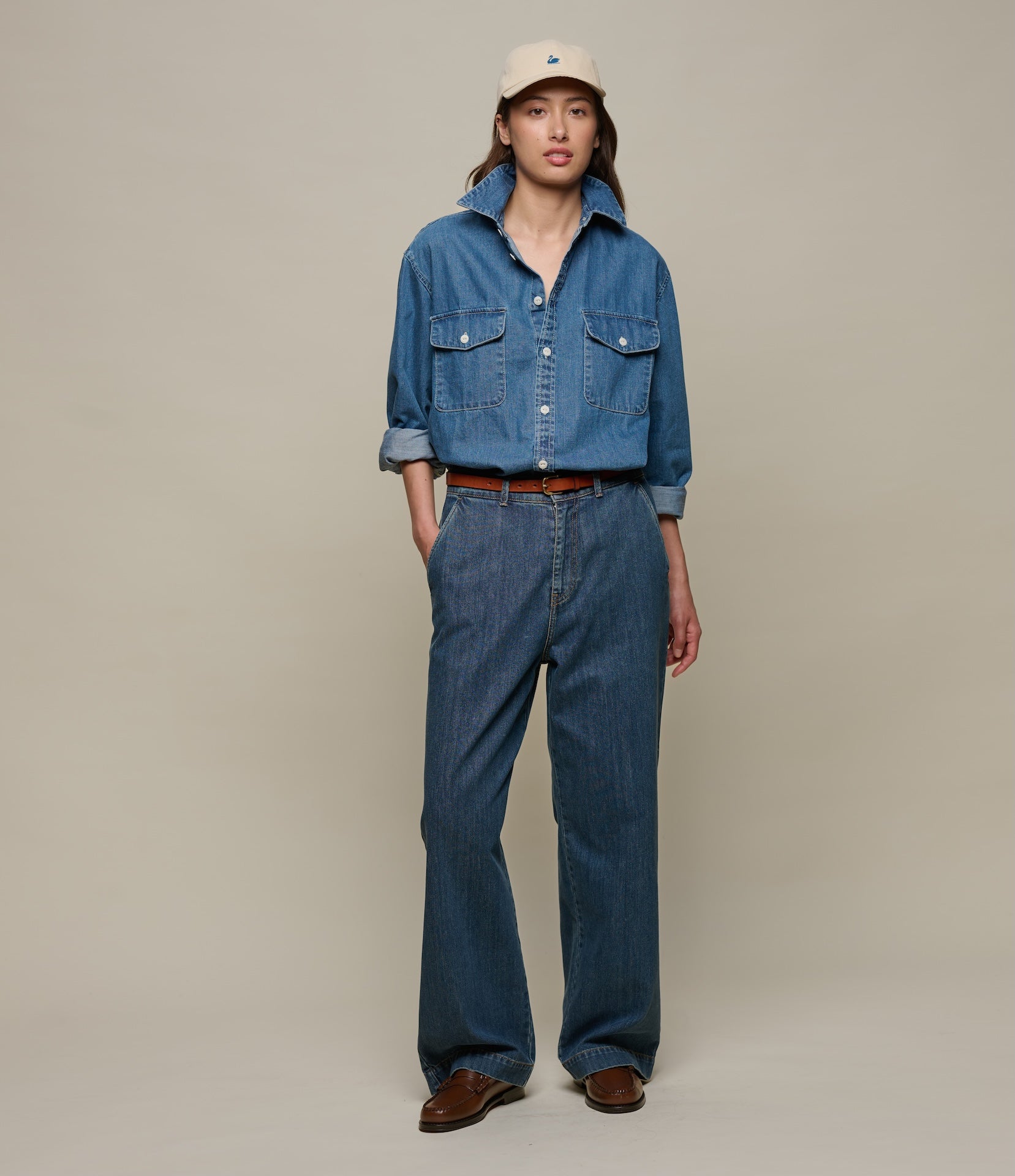 Person wearing a blue denim outfit with a beige cap on a plain background