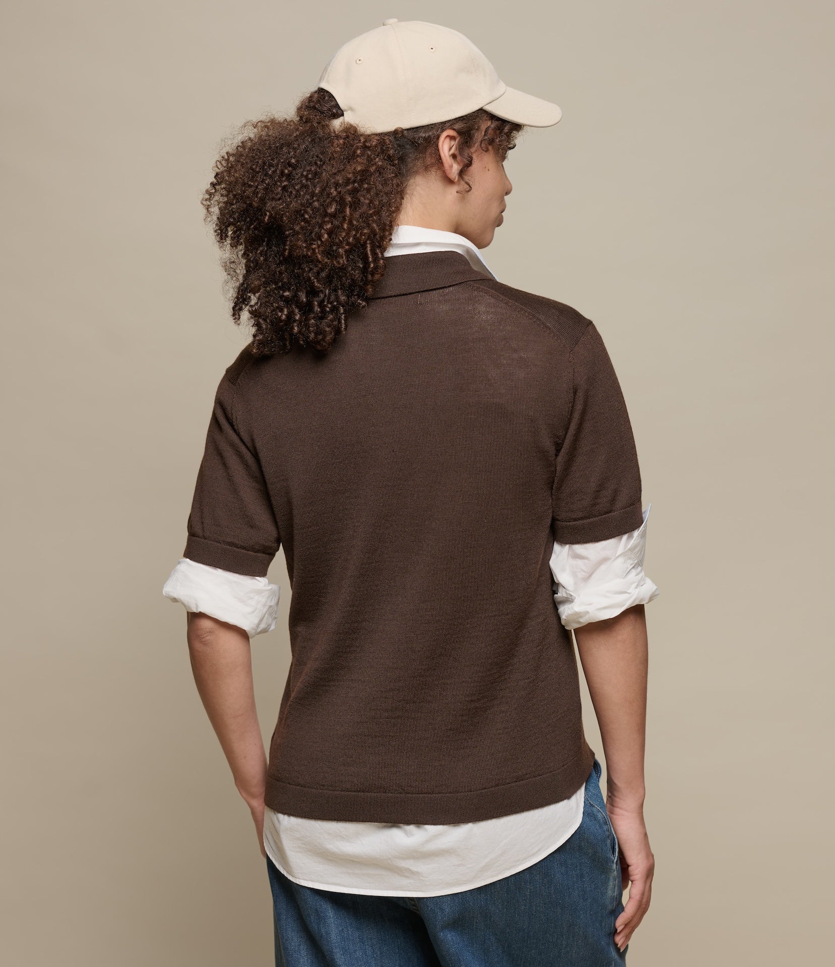 Person wearing a brown knitted polo shirt with rolled-up sleeves and a beige cap on a beige background
