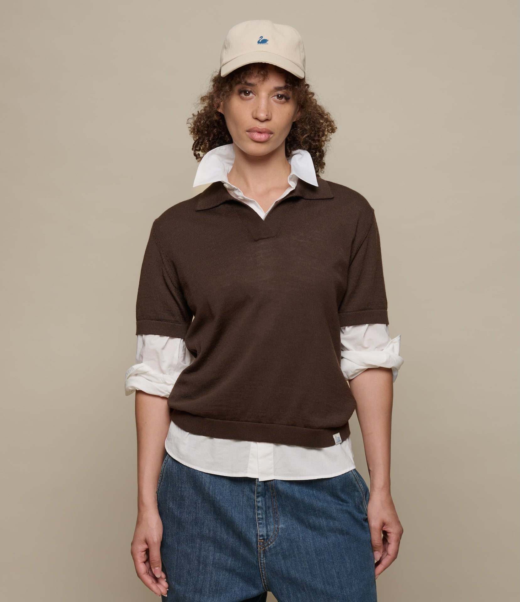 Woman wearing a brown knitted polo shirt over a white shirt with rolled-up sleeves, blue jeans, and a beige cap on a beige background.