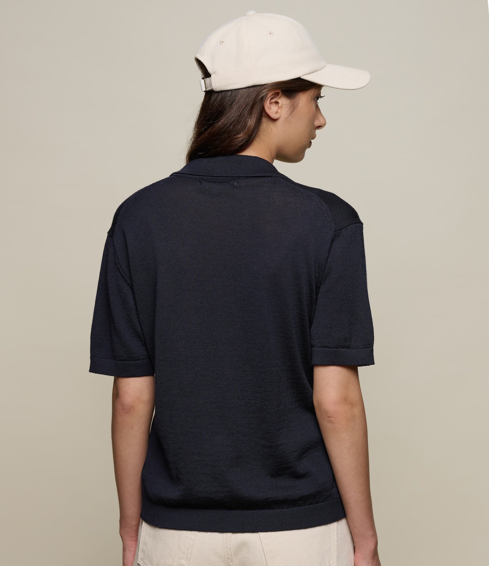 Person wearing a navy knitted polo shirt and beige cap on a plain background