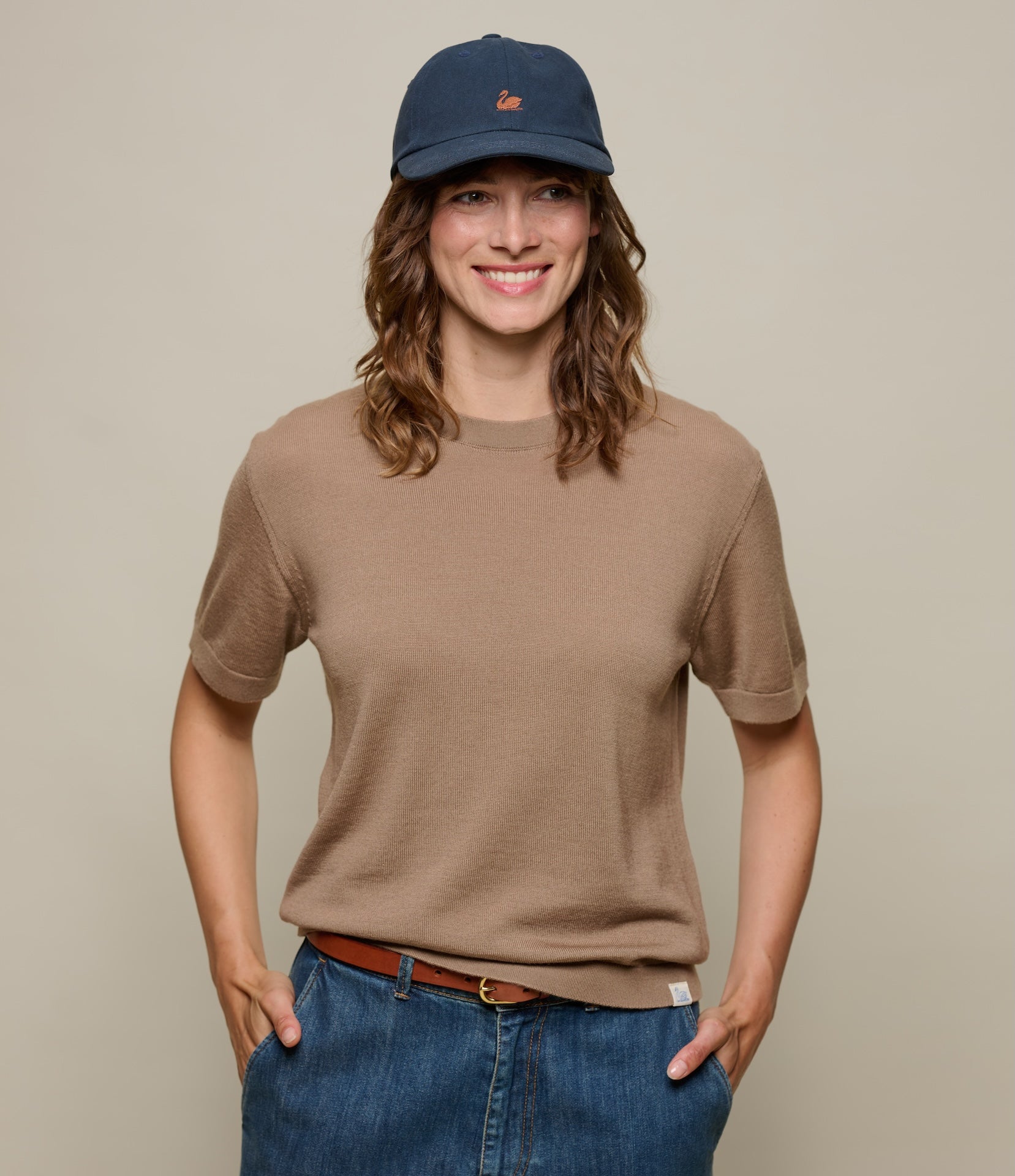 Person wearing a brown knitted t-shirt and blue jeans with a navy cap on a beige background