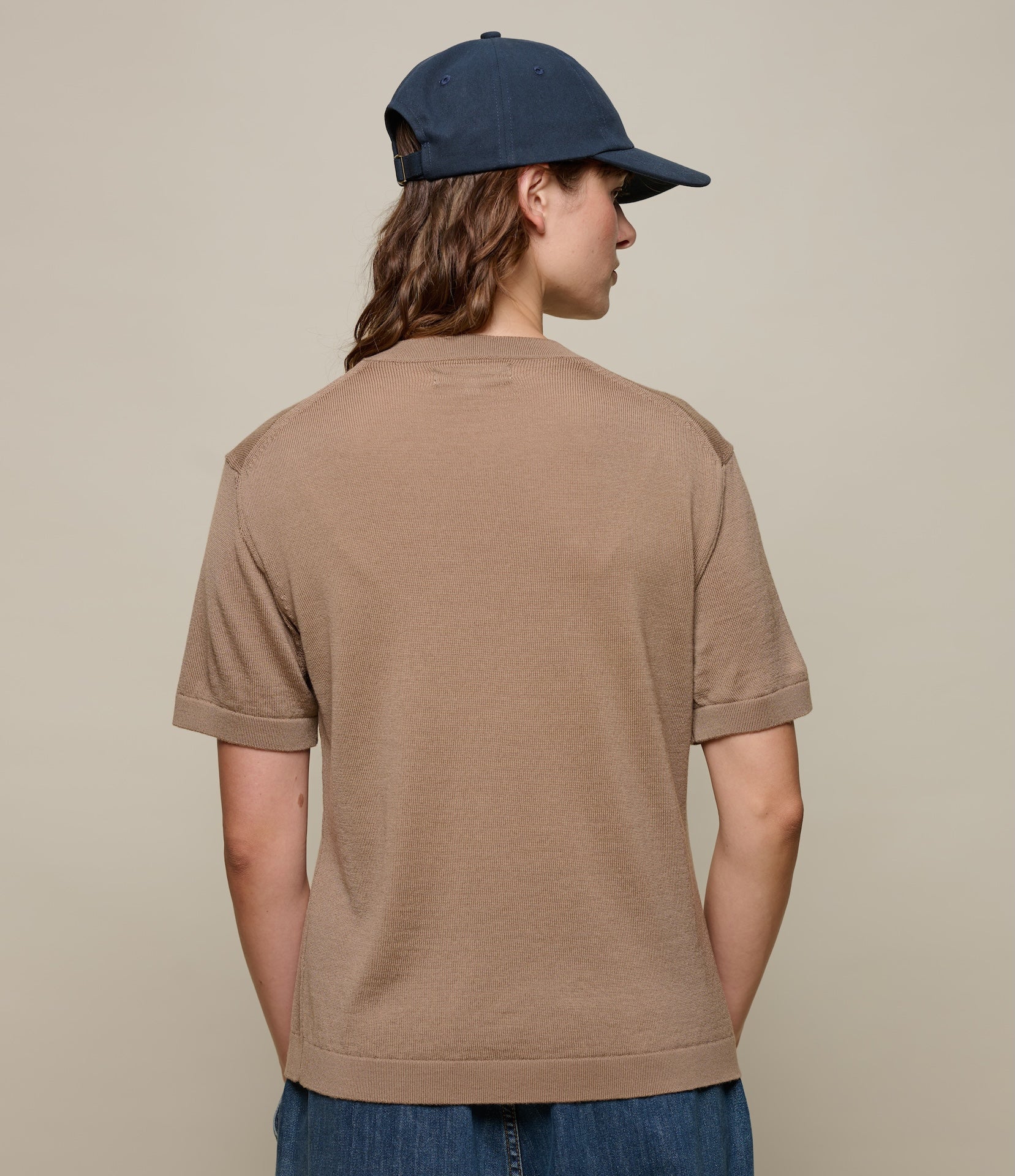 Person wearing a brown knitted t-shirt and navy cap on a beige background