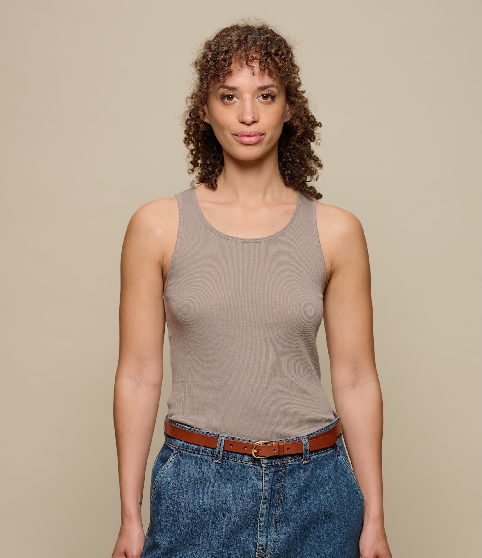 Woman wearing a brown ribbed tank top and blue jeans against a beige background