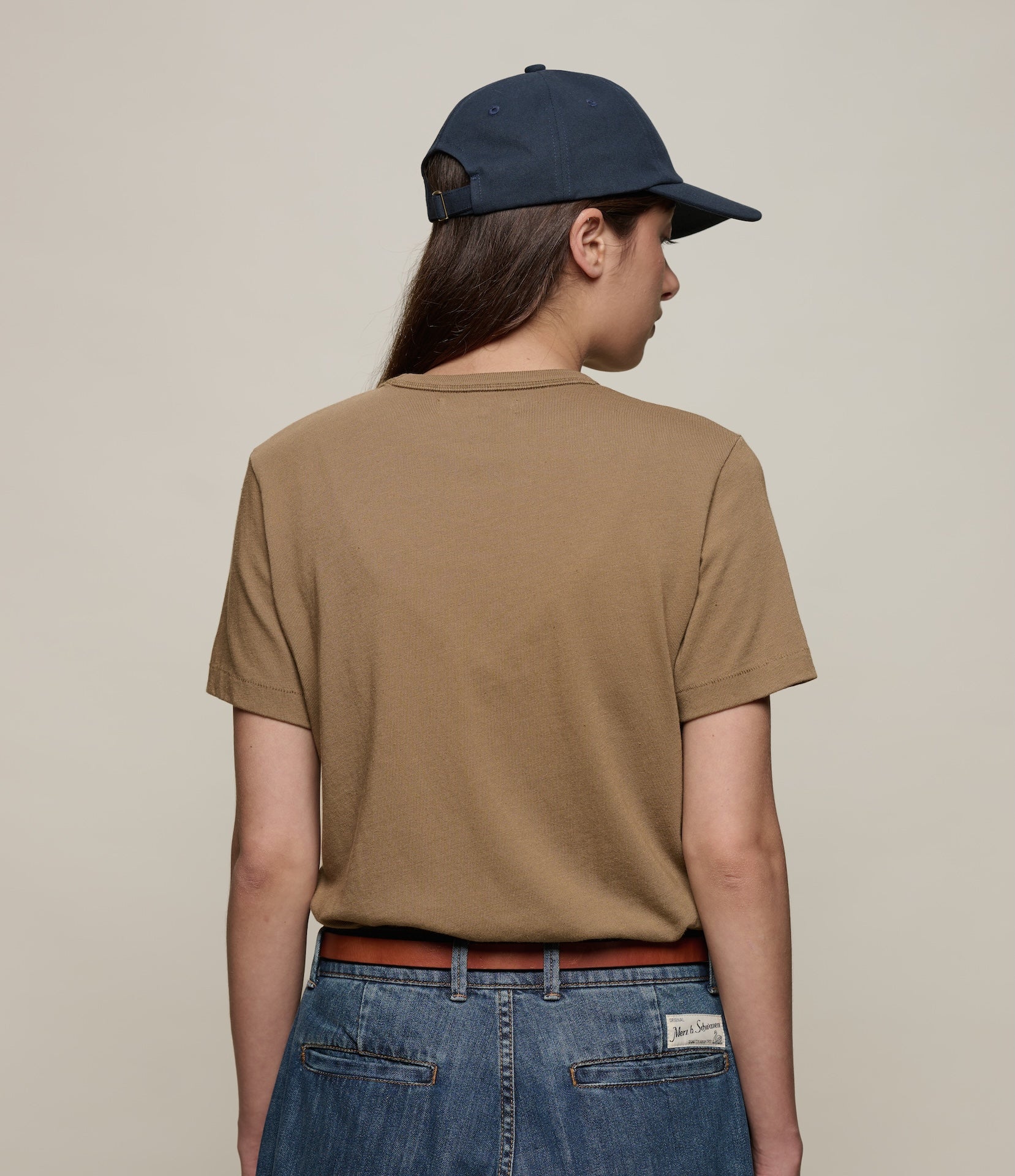 Person wearing a brown t-shirt, blue jeans, and a navy cap on a beige background