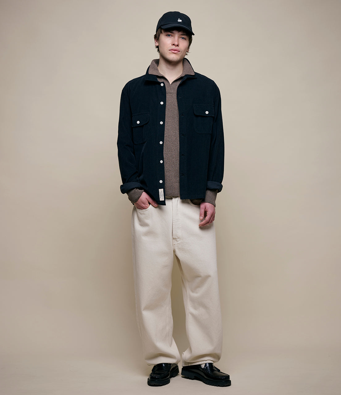 Person wearing a black jacket, beige sweater, white pants, and black shoes on a beige background